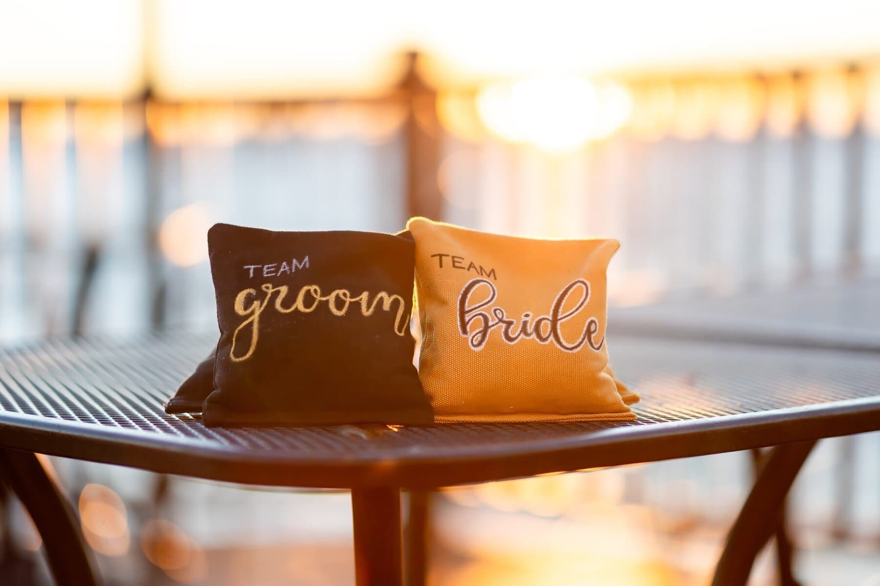 Wedding at the Newport Naval Station Officers' Club 79 Two small pillows labeled "Team Groom" and "Team Bride" sit on a metal table at an Officers' Club Wedding with sunlight in the background.