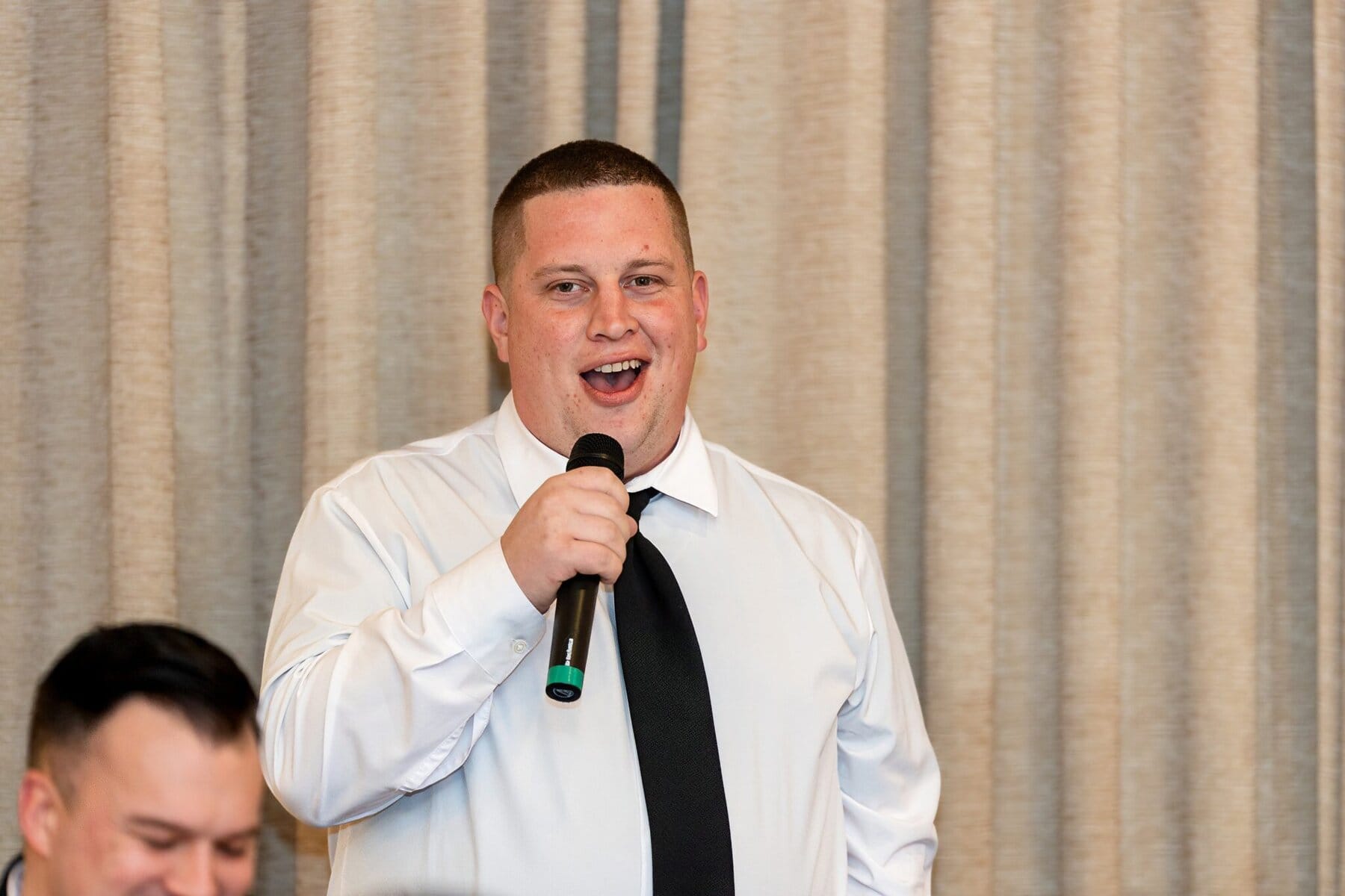 Wedding at the Newport Naval Station Officers' Club 81 Man in a white shirt and black tie speaks into a microphone at a wedding in front of beige curtains at Newport Naval Station.