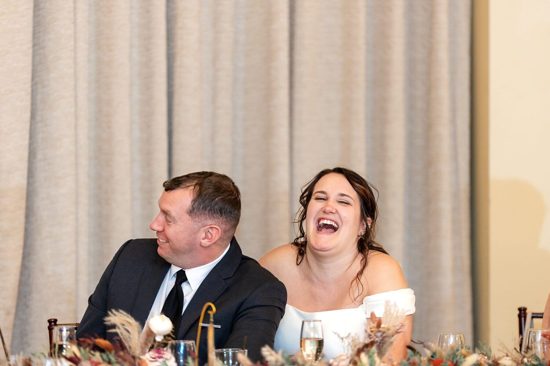 Wedding at the Newport Naval Station Officers' Club 82 At a wedding at the Officers' Club, a man in a suit and a laughing woman in white sit together at the table.