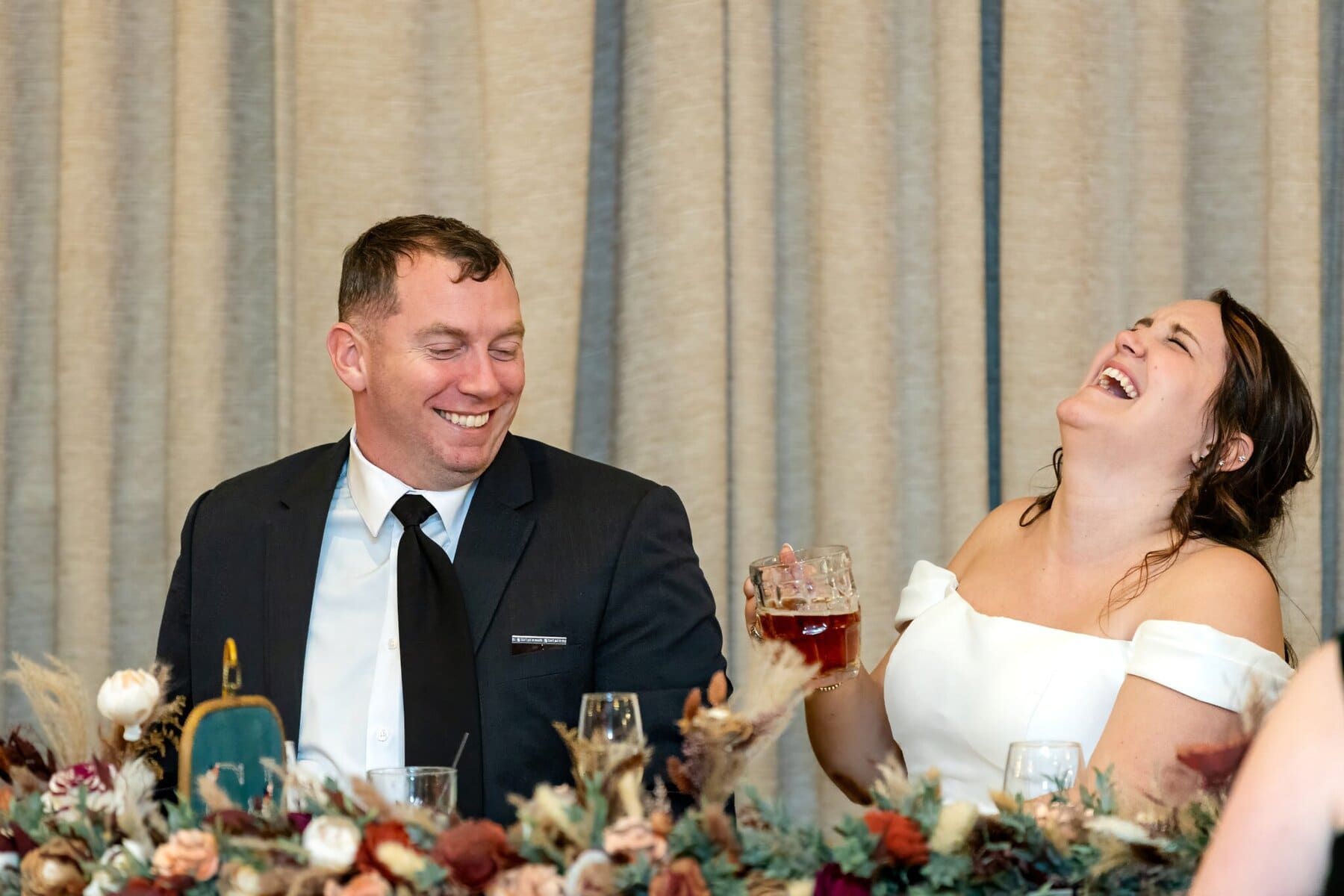 Wedding at the Newport Naval Station Officers' Club 83 A man in a suit and a woman in a white dress laugh together at their wedding table, drinks in hand, beige curtains behind.