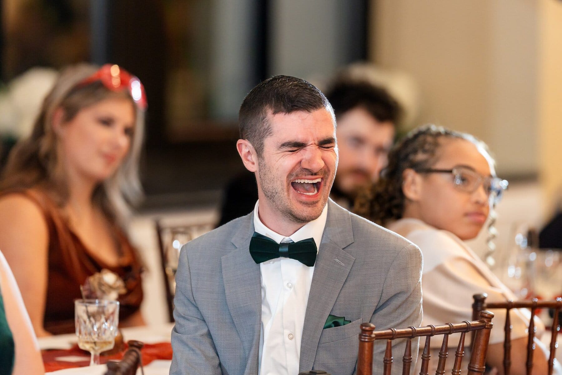 Wedding at the Newport Naval Station Officers' Club 86 A man in a gray suit and green bow tie laughs at a wedding at the Officers' Club, with other guests blurred in the background.