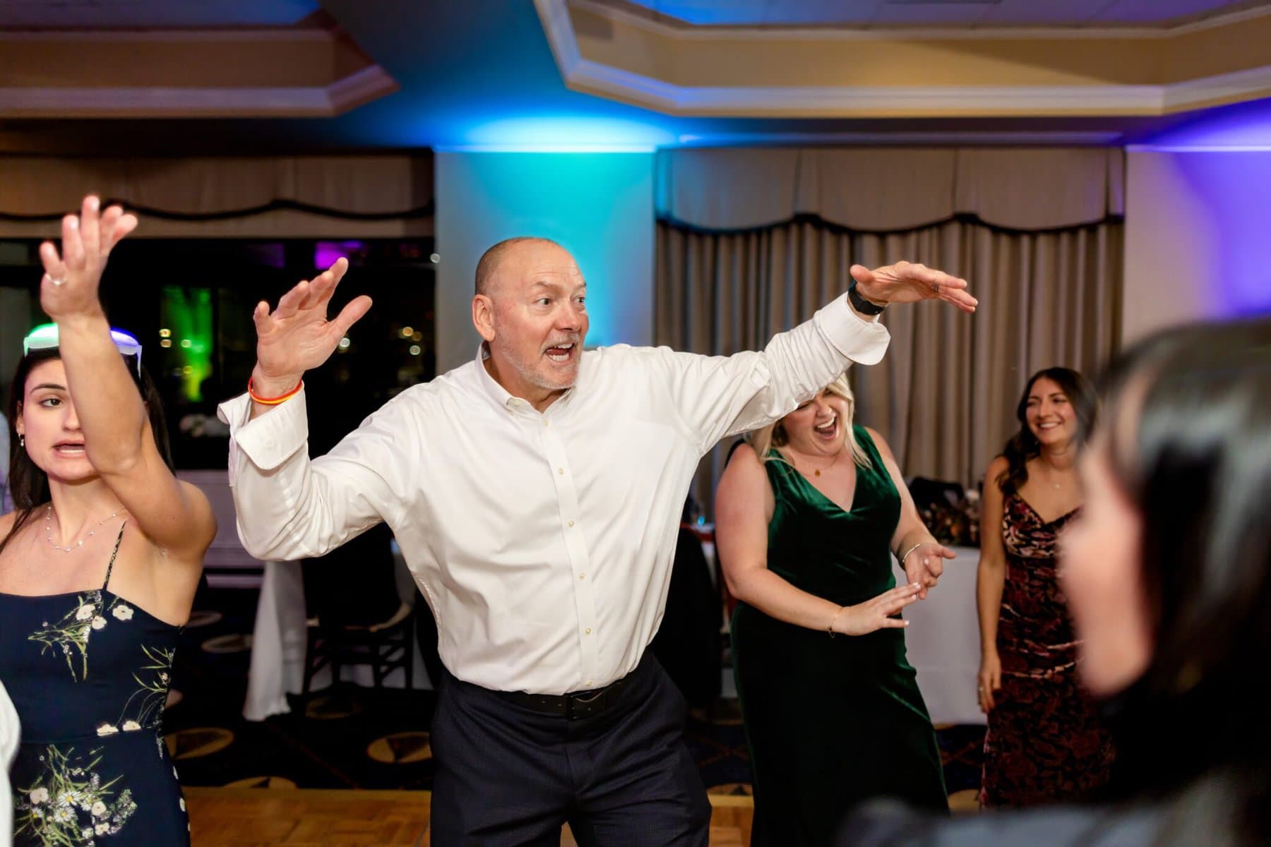 Wedding at the Newport Naval Station Officers' Club 98 A man in a white shirt dances enthusiastically at an indoor Newport wedding, surrounded by smiling people in formal attire.