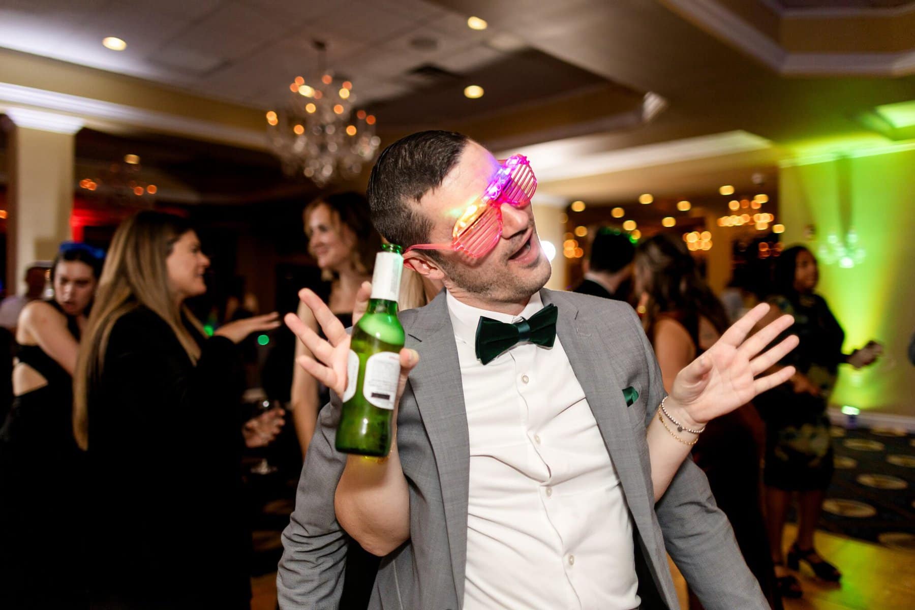 Wedding at the Newport Naval Station Officers' Club 101 A man in a suit and bow tie wears light-up glasses, holding a beer while dancing at a Newport Naval Station wedding's Officers' Club party.