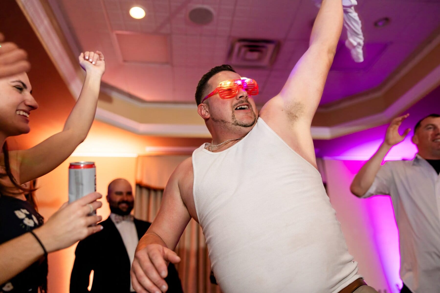 Wedding at the Newport Naval Station Officers' Club 105 A man in a white tank top and light-up glasses dances at a wedding at the Officers' Club, Newport Naval Station, surrounded by cheering guests.