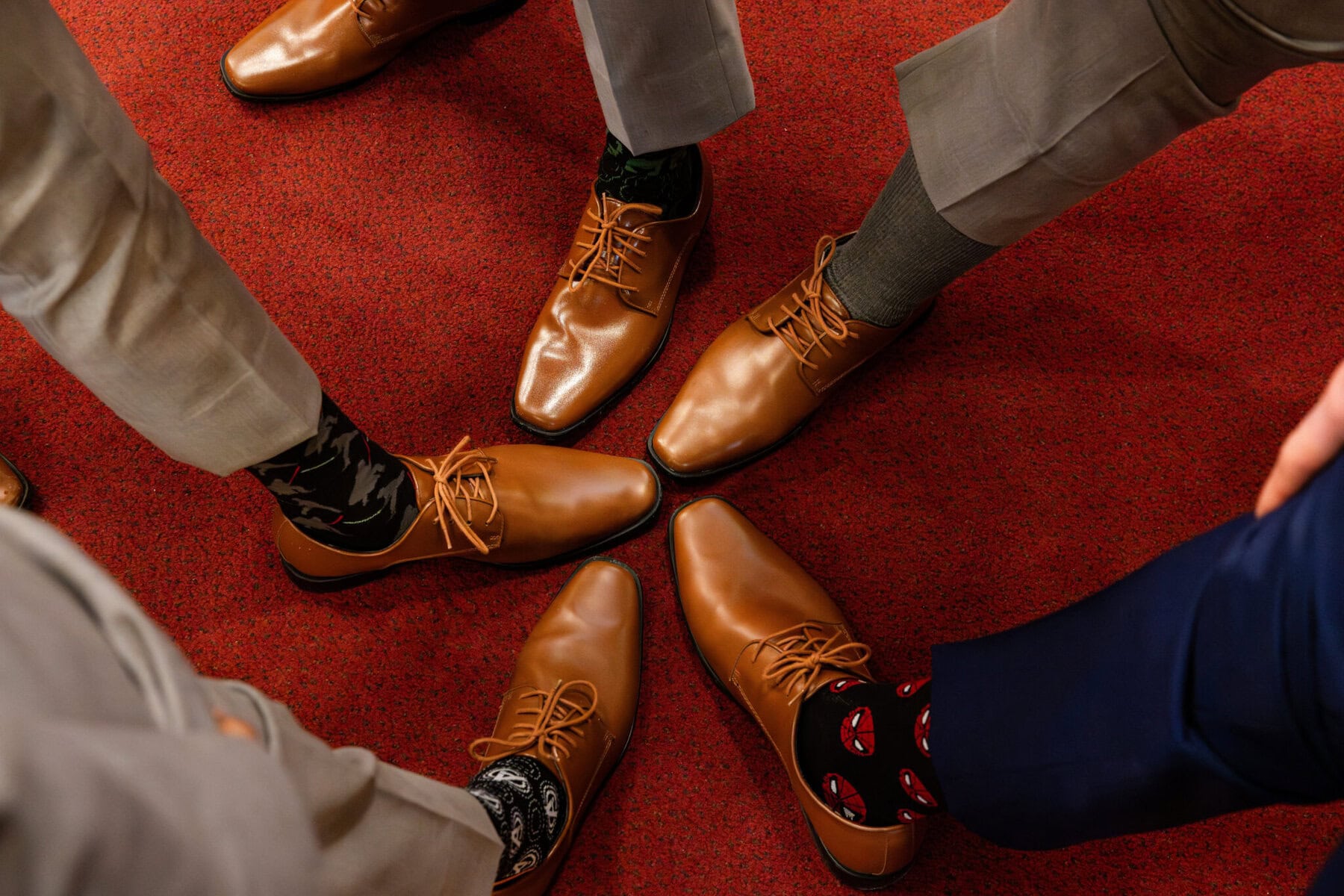 Wedding At the Transfiguration of the Lord Parish 18 Four people in tan dress shoes and colorful socks stand on a red carpet at a wedding, feet pointed toward the center.