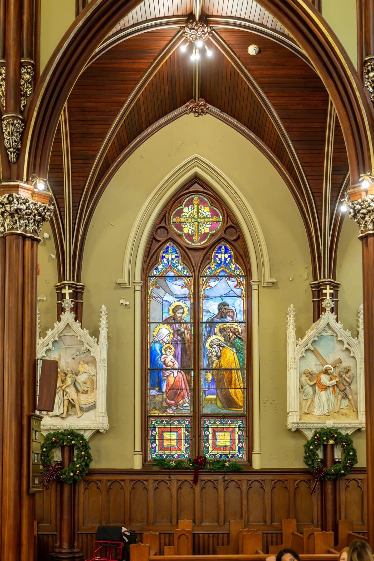 Wedding At the Transfiguration of the Lord Parish 16 Stained glass window of the Transfiguration of the Lord in a church above wooden pews, flanked by statues and wreaths, beneath a vaulted ceiling.