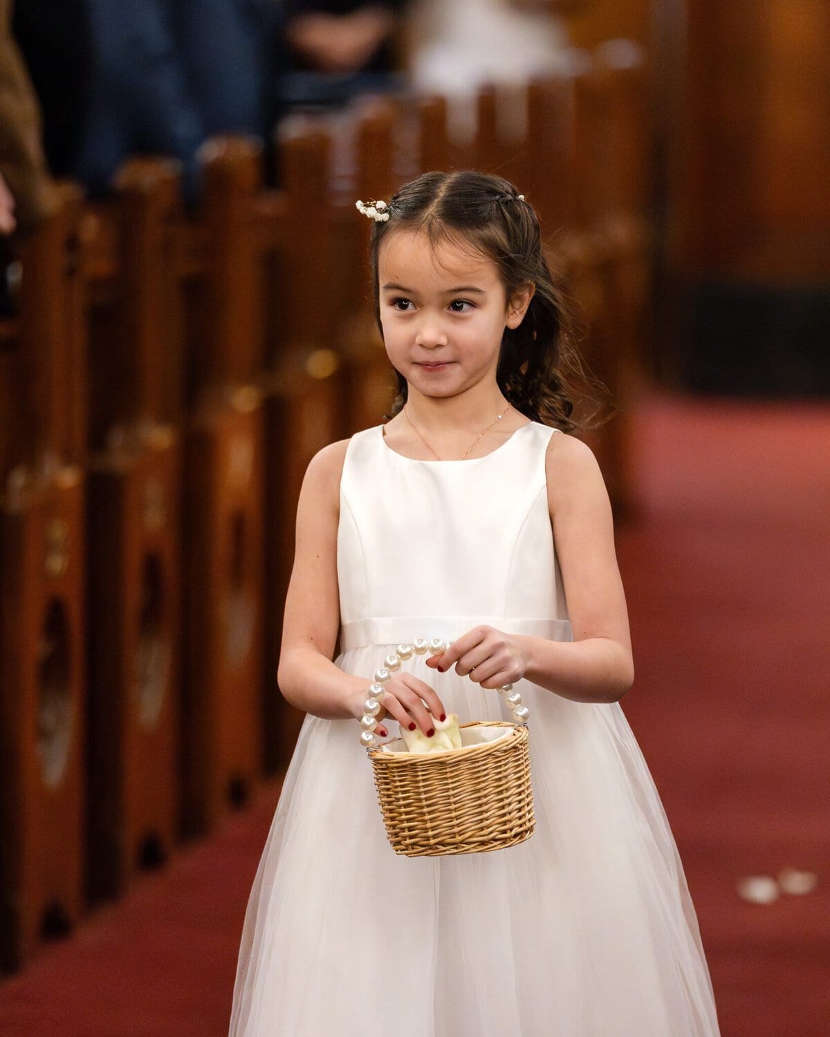Wedding At the Transfiguration of the Lord Parish 25 A young girl in a white dress walks down an aisle with a wicker basket, likely during a wedding at Transfiguration of the Lord Parish.