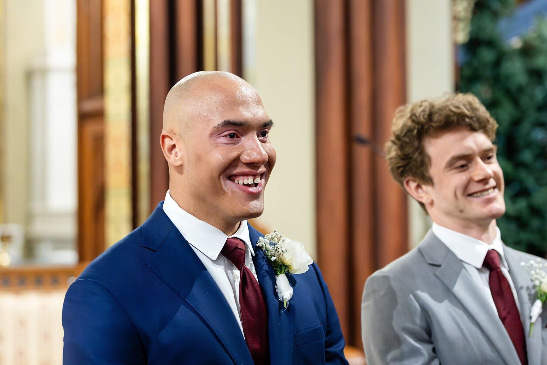 Wedding At the Transfiguration of the Lord Parish 30 Two men in suits with boutonnieres smile indoors at a Transfiguration of the Lord Parish wedding.