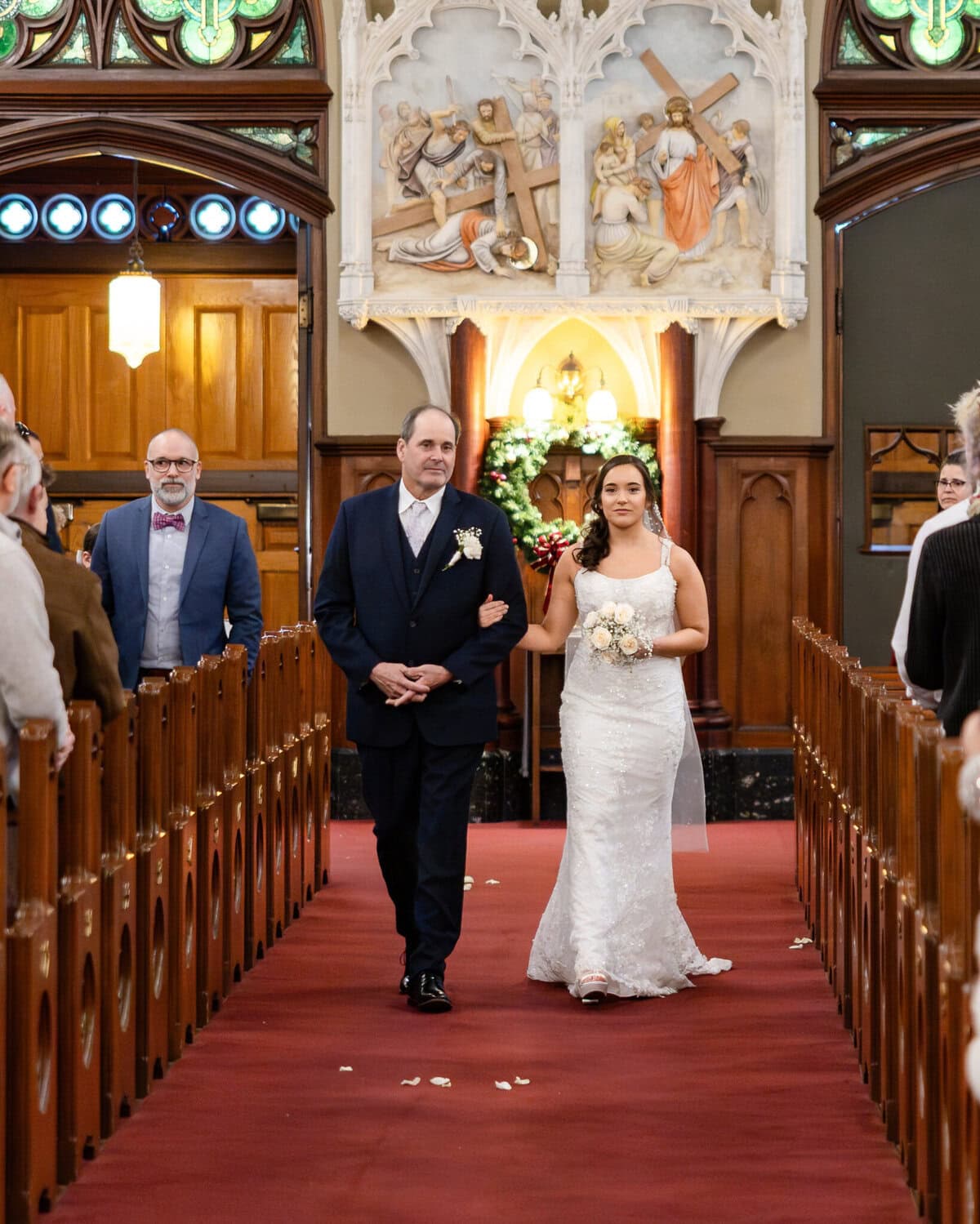 Wedding At the Transfiguration of the Lord Parish 31 A bride in white walks down the aisle at Transfiguration of the Lord Parish, wedding guests seated on ornate wooden pews.
