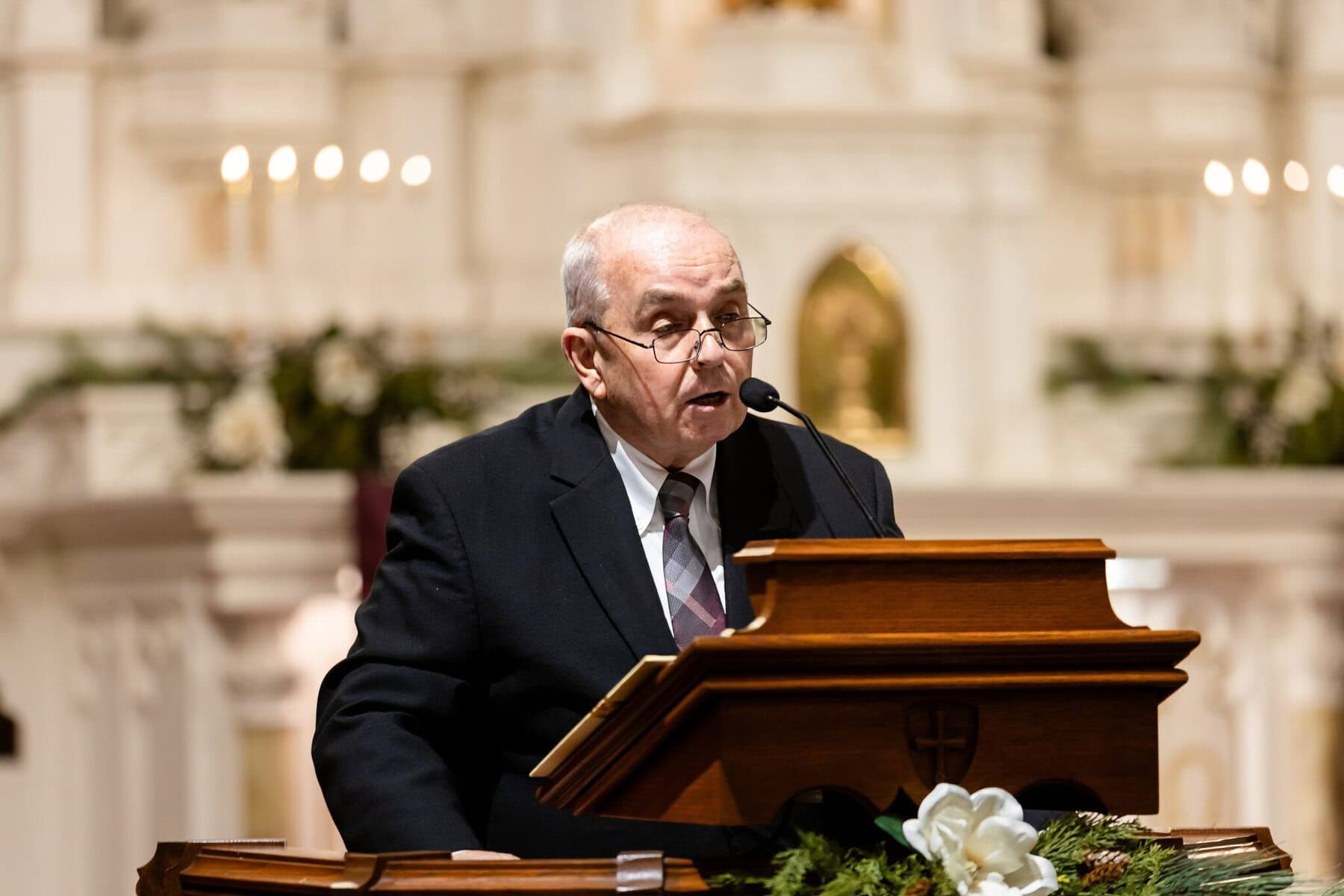Wedding At the Transfiguration of the Lord Parish 34 An older man in a suit speaks at a wooden podium in Transfiguration of the Lord Parish, with candles and floral wedding decorations behind him.