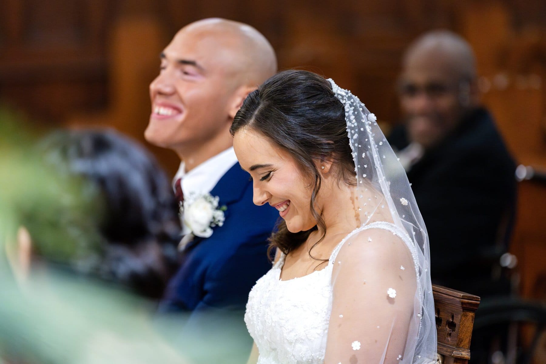 Wedding At the Transfiguration of the Lord Parish 38 A bride and groom sit side by side, smiling during a parish wedding at Transfiguration of the Lord Parish, with guests in the background.
