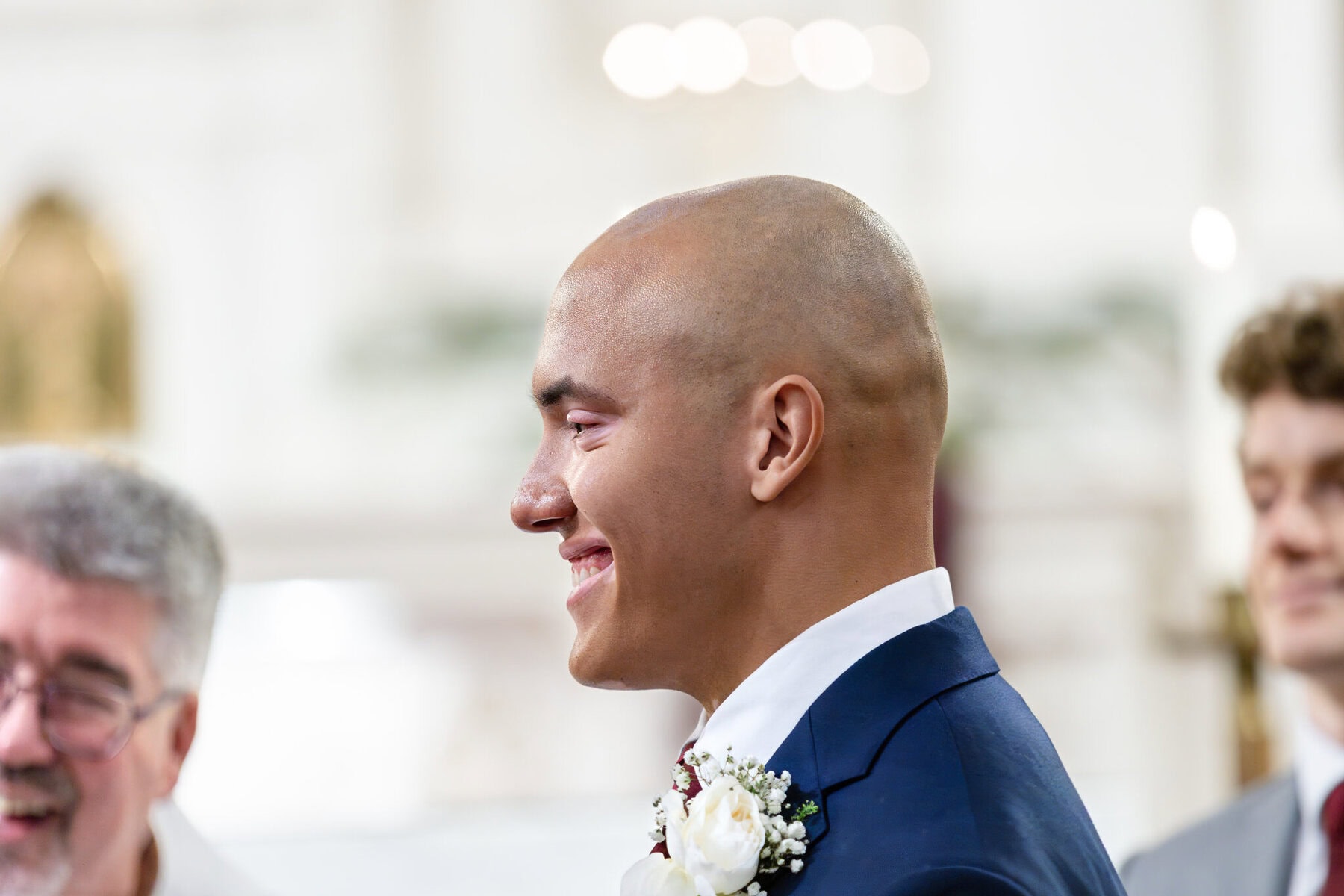 Wedding At the Transfiguration of the Lord Parish 42 A man in a suit with a boutonniere smiles, standing indoors at a Transfiguration of the Lord Parish formal event.