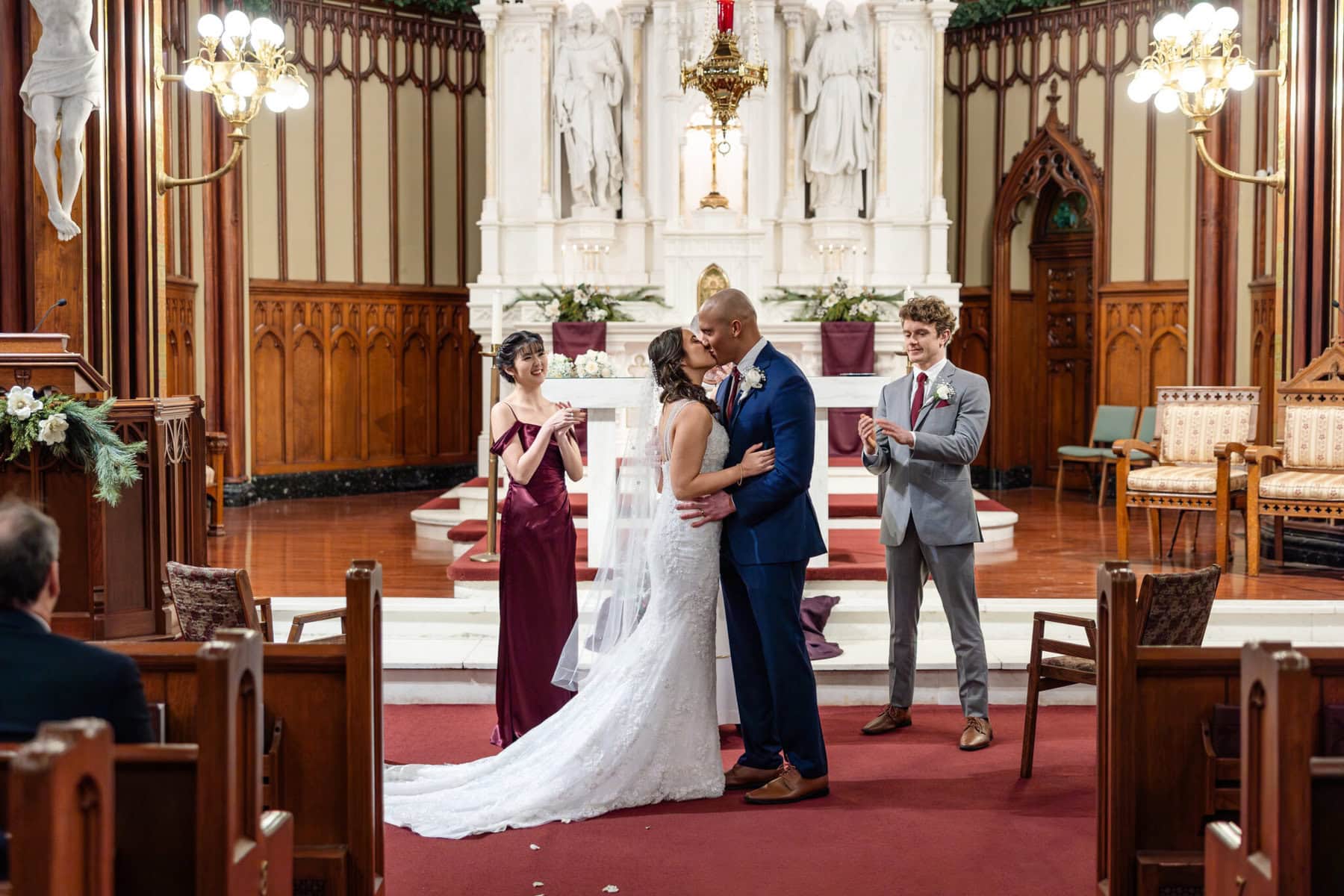 Wedding At the Transfiguration of the Lord Parish 45 A bride and groom kiss at the altar during a wedding at Transfiguration of the Lord Parish as attendants stand and applaud.