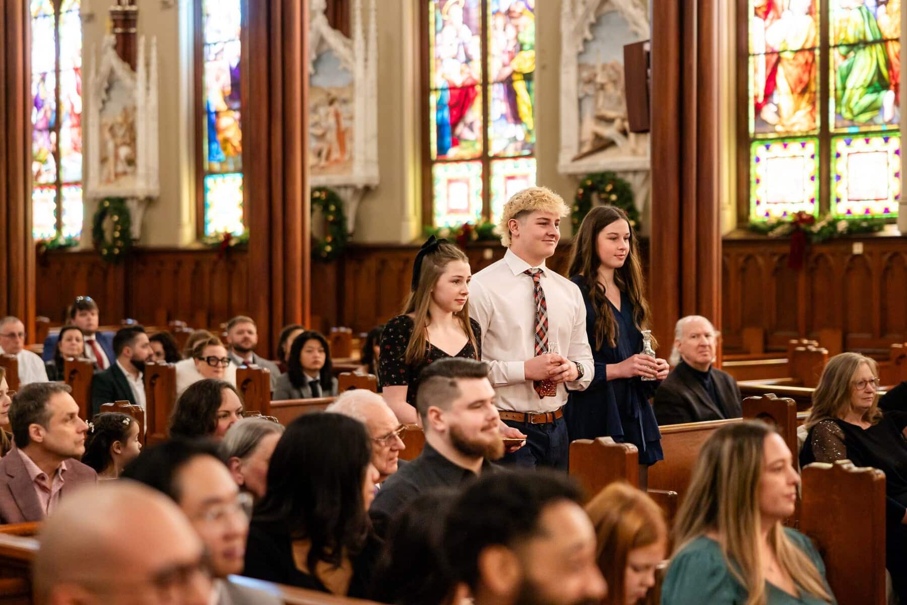 Wedding At the Transfiguration of the Lord Parish 47 Three young people stand in the aisle of Transfiguration of the Lord Parish as others sit in pews, stained glass windows glowing behind them.