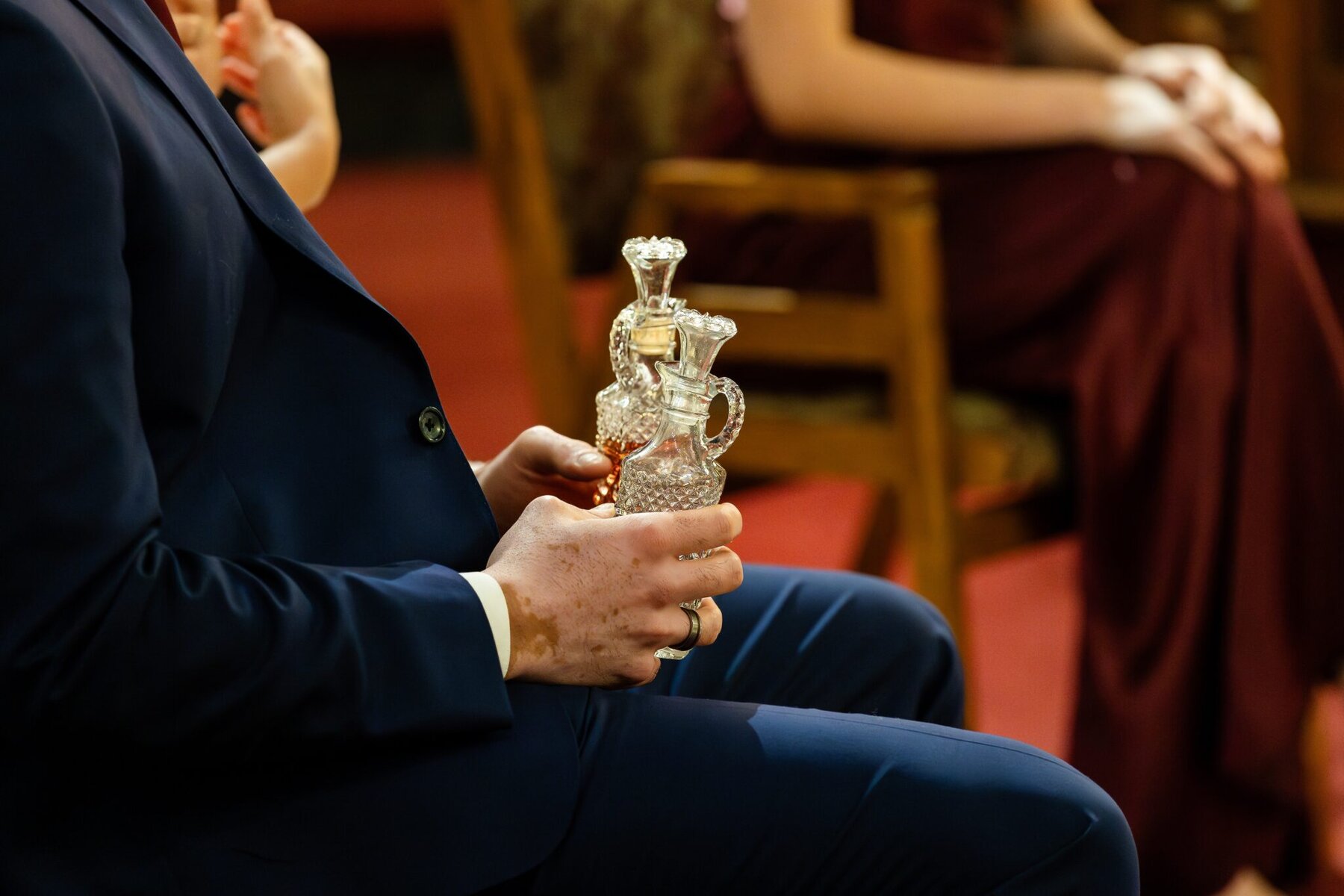 Wedding At the Transfiguration of the Lord Parish 49 A person in a blue suit sits holding ornate glass decanters at Transfiguration of the Lord Parish; others are seated nearby.