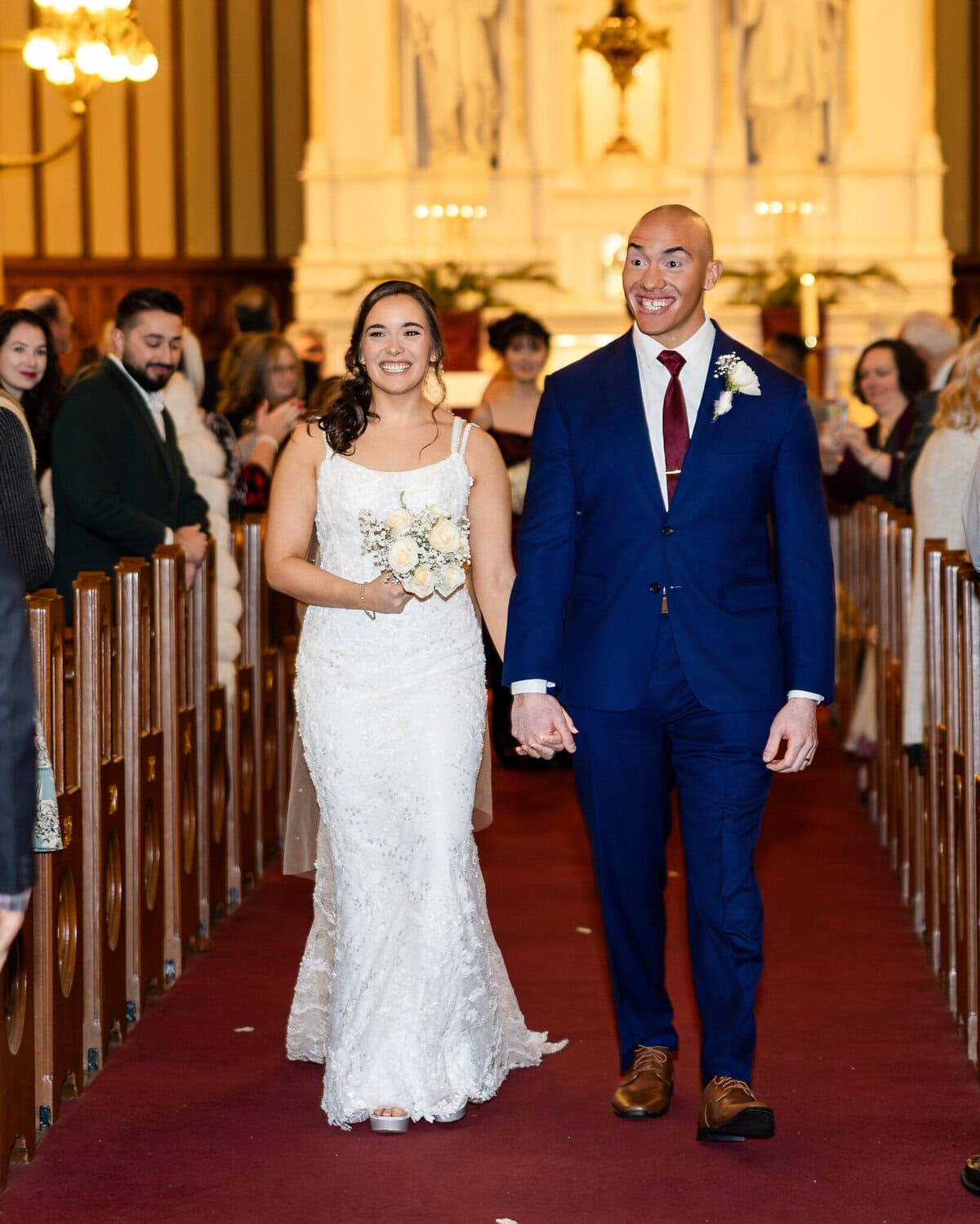 Wedding At the Transfiguration of the Lord Parish 50 A bride and groom walk down the aisle at Transfiguration of the Lord Parish, holding hands, with guests seated on both sides.