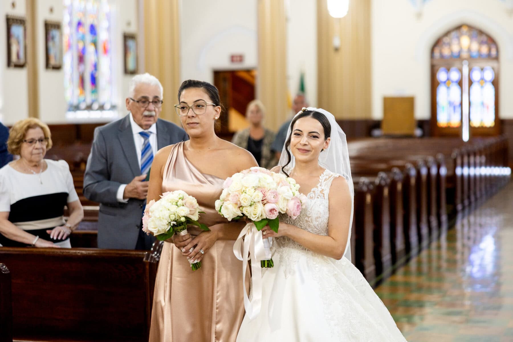 Wedding At Santo Christo Parish in Fall River 20 A bride in a white gown walks down the Santo Christo Parish aisle in Fall River, with her bridesmaid; guests watch this wedding from the pews.