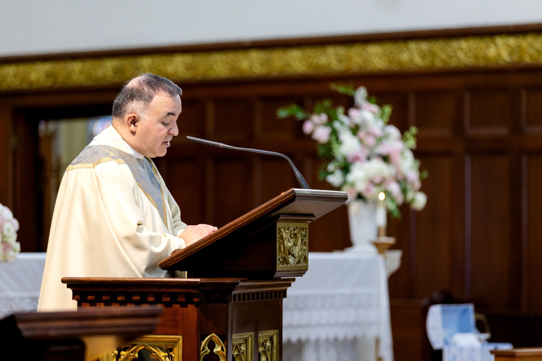 Wedding At Santo Christo Parish in Fall River 35 A man in religious robes stands at a lectern, speaking in Santo Christo Parish, a church in Fall River adorned with flowers and wooden paneling.