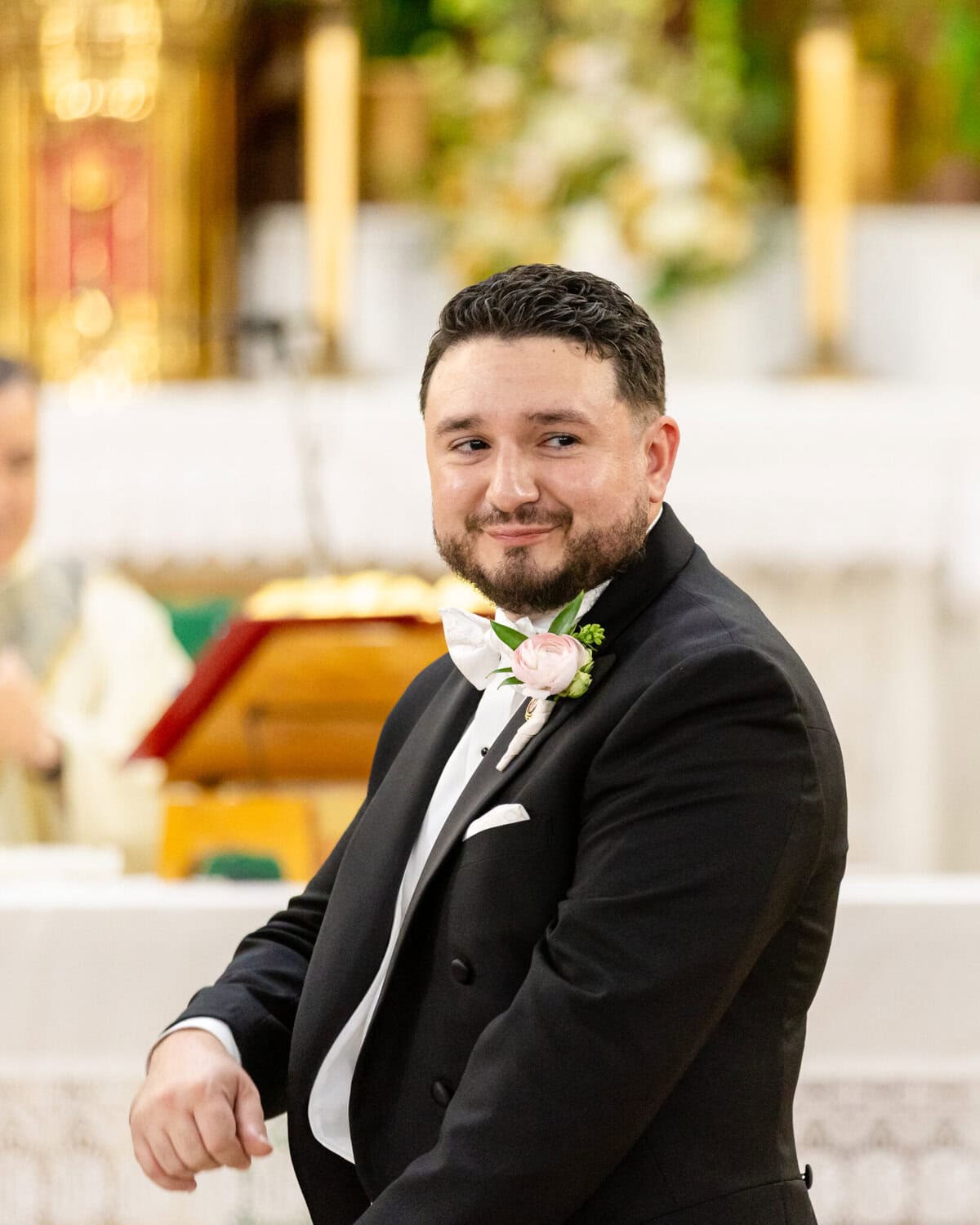 Wedding At Santo Christo Parish in Fall River 45 A man in a tuxedo with a boutonniere smiles indoors, with blurred Santo Christo Parish wedding decor in Fall River behind him.