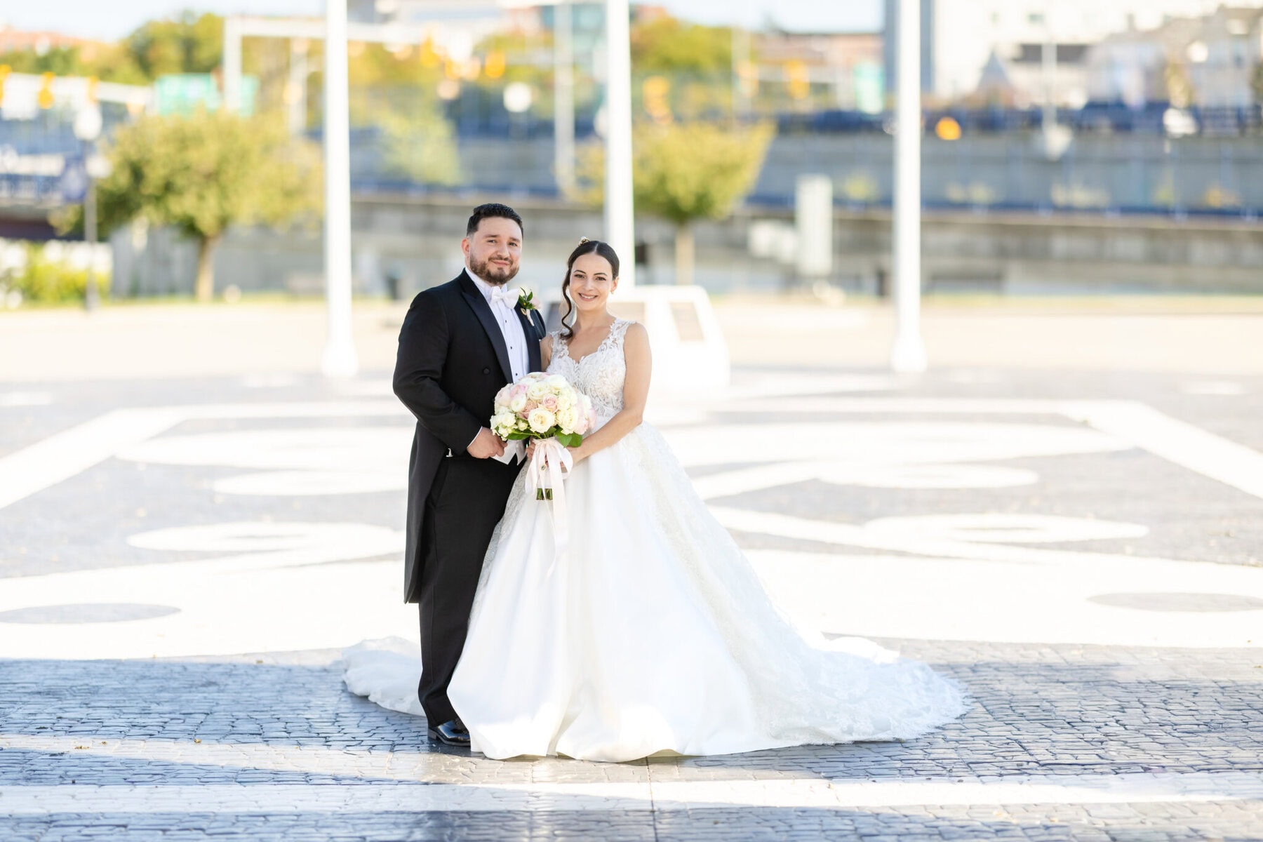 Wedding Photos at City Gates Plaza in Fall River 2 A bride and groom stand outdoors in wedding attire at City Gates Plaza, smiling and holding a bouquet of flowers on a bright day.