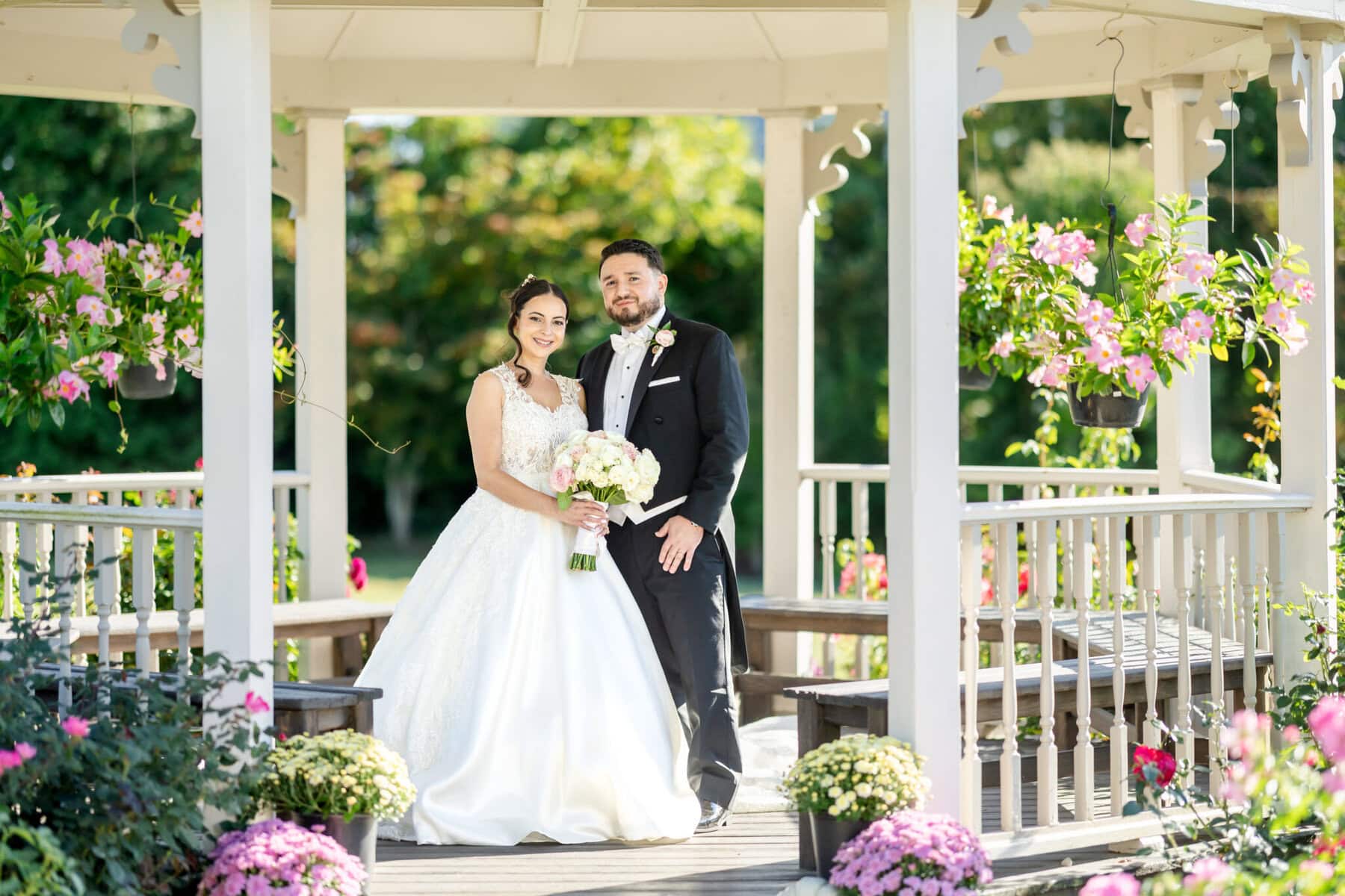 Wedding Photos at Roseland Nursery 5 A bride and groom stand together under a gazebo at Roseland Nursery, both smiling for their wedding photos amid lush flowers and greenery.