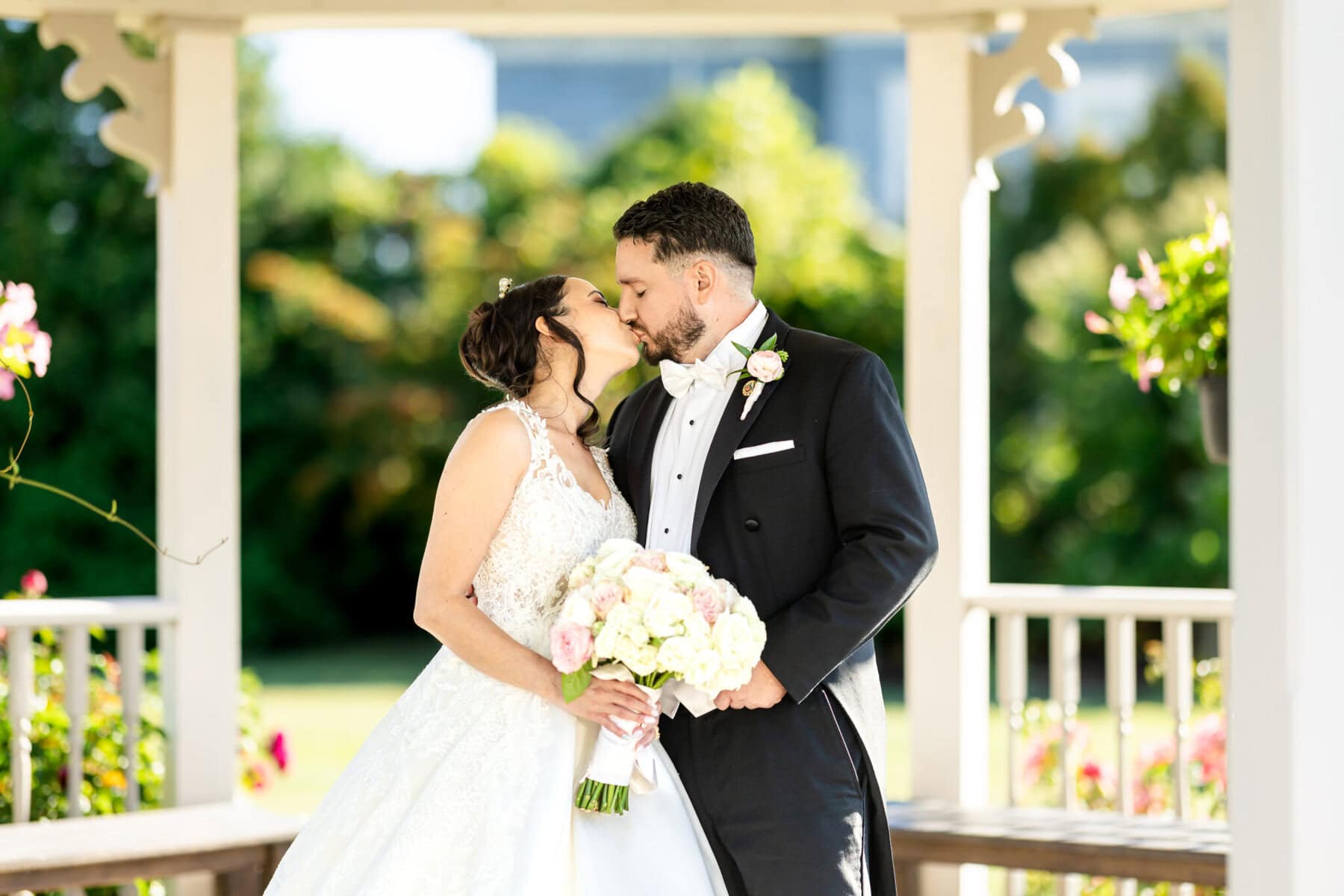 Wedding Photos at Roseland Nursery 3 A bride and groom in formal attire kiss under a gazebo at Roseland Nursery, surrounded by lush greenery—perfect for wedding photography.