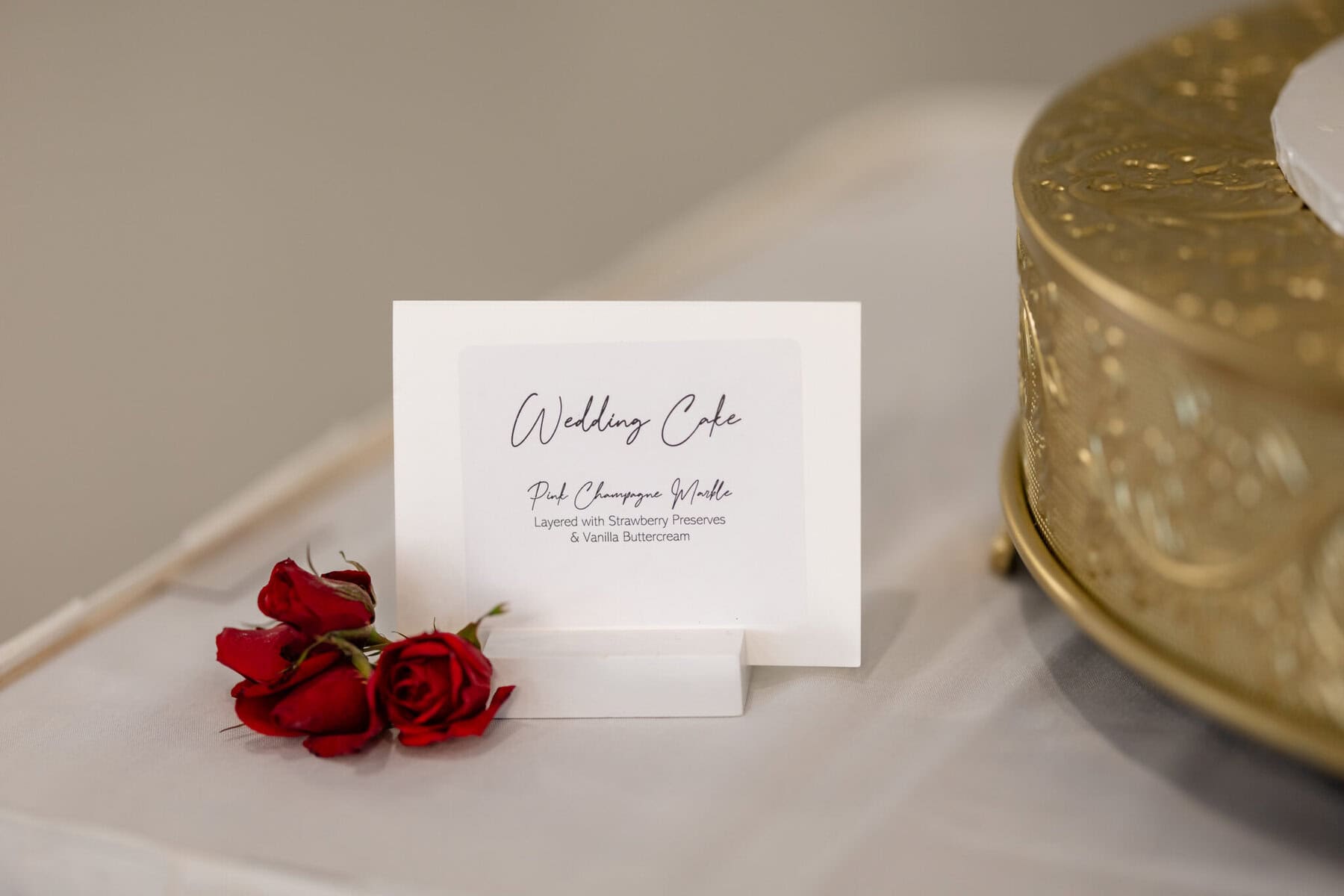 Wedding Reception at The Century House in Acushnet 6 A wedding cake label with two red roses beside it, on a white table at The Century House wedding reception in Acushnet.