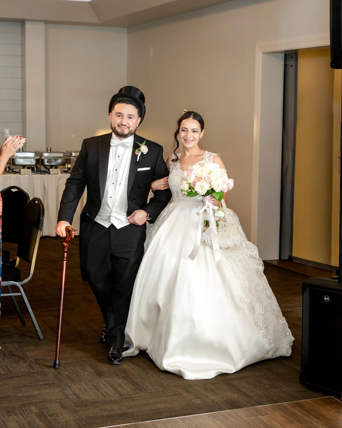 Wedding Reception at The Century House in Acushnet 12 A bride and groom walk arm in arm indoors at The Century House; the groom wears a tuxedo and top hat, the bride holds a bouquet of flowers.