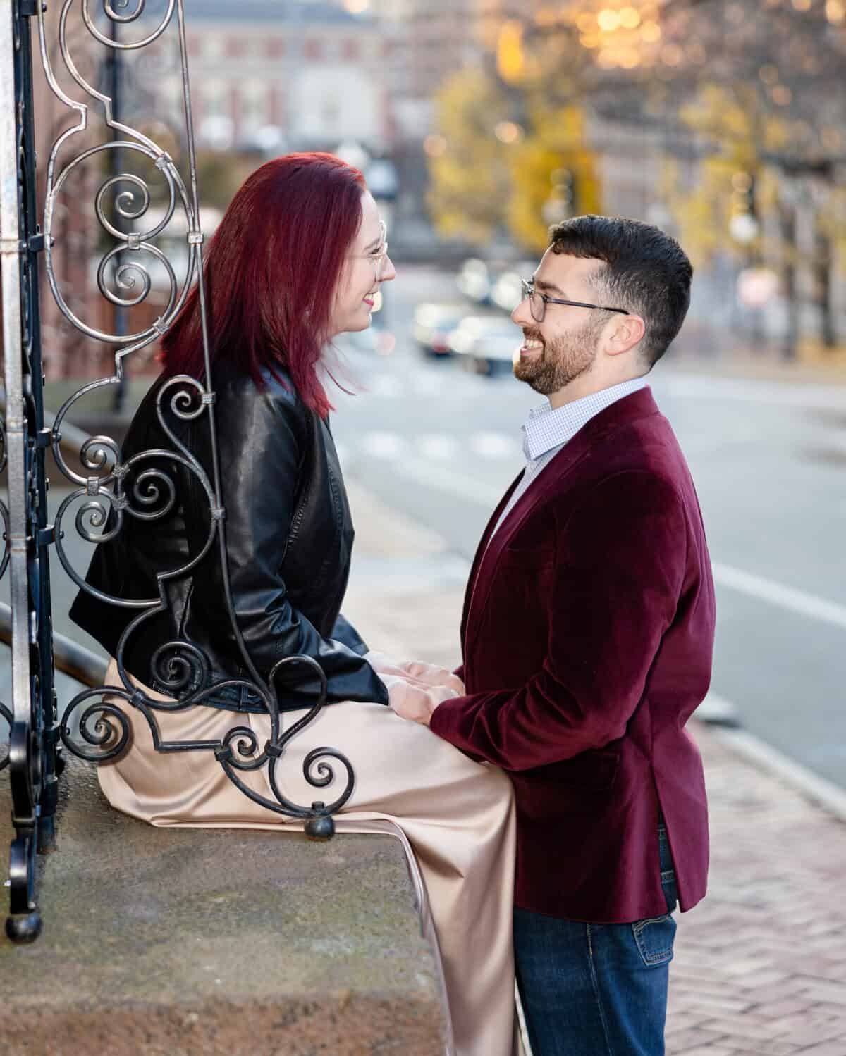 Downtown Providence Engagement Session 4 Brittany and Greg smile at each other on a city sidewalk during their Downtown Providence engagement session; she sits in gold and black, while he stands in a burgundy blazer, holding her hands.
