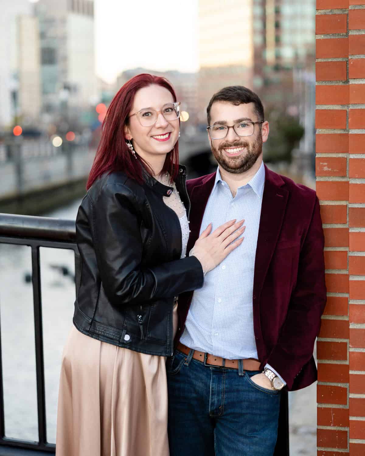 Downtown Providence Engagement Session 5 Brittany and Greg, both in glasses, smile as they pose near a brick wall and cityscape during their engagement session in downtown Providence.