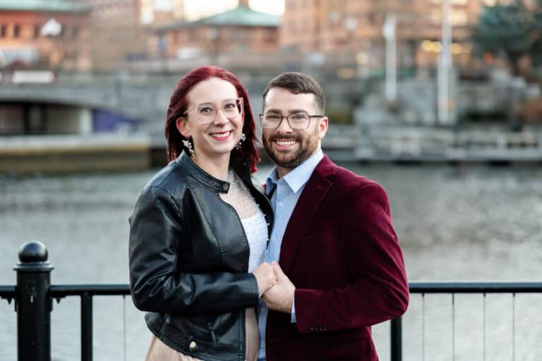 Brittany and Greg share a joyful moment during their engagement session by a riverside railing in downtown Providence, dressed stylishly and smiling together.