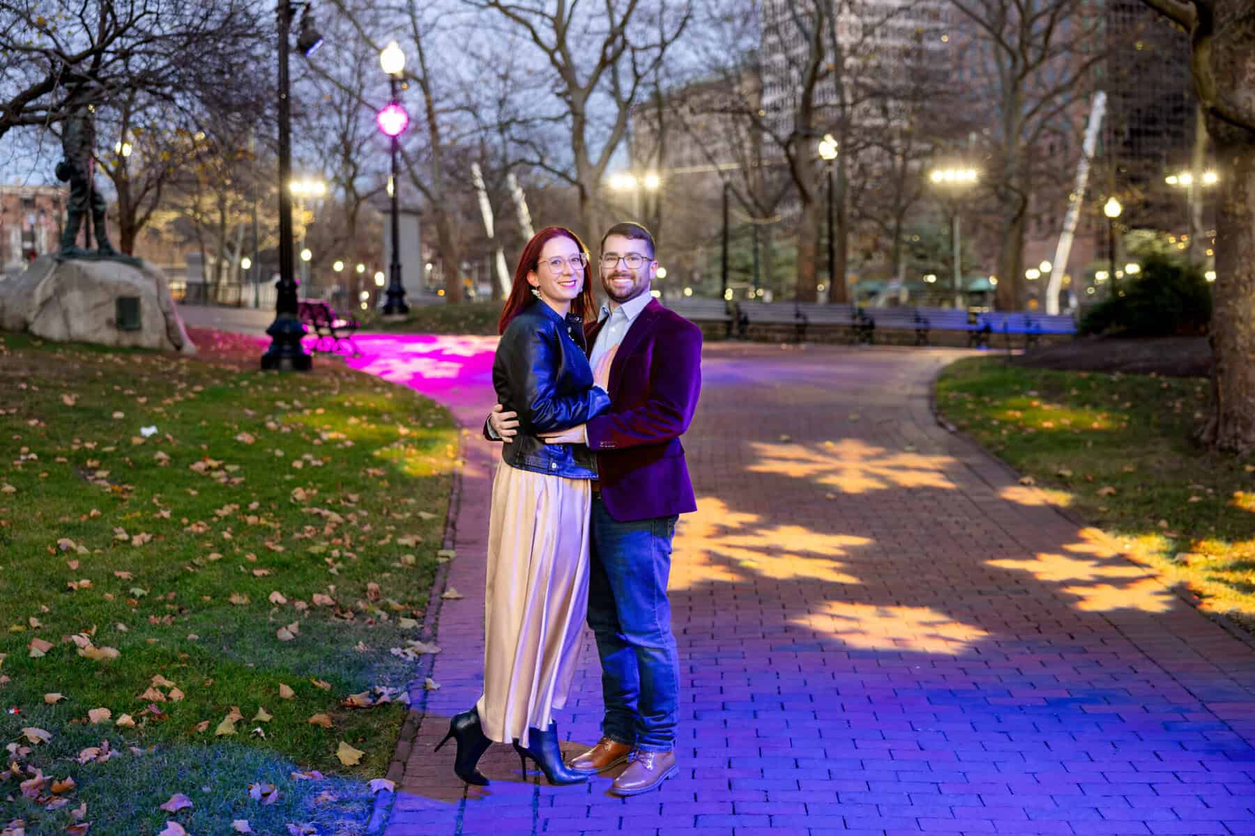 Downtown Providence Engagement Session 26 Brittany and Greg stand smiling and embracing on a brick path in Downtown Providence at dusk, surrounded by trees and festive snowflake lights projected on the ground.