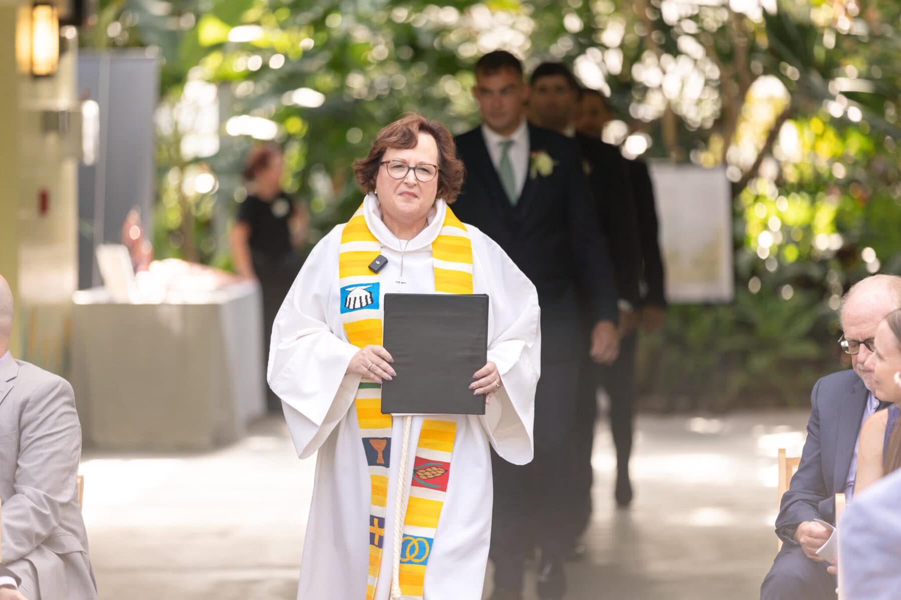 Stunning Wedding at the Roger Williams Botanical Gardens 15 A woman in a white robe with a colorful stole leads a wedding procession at Roger Williams Botanical Gardens, holding a folder in a bright indoor space.