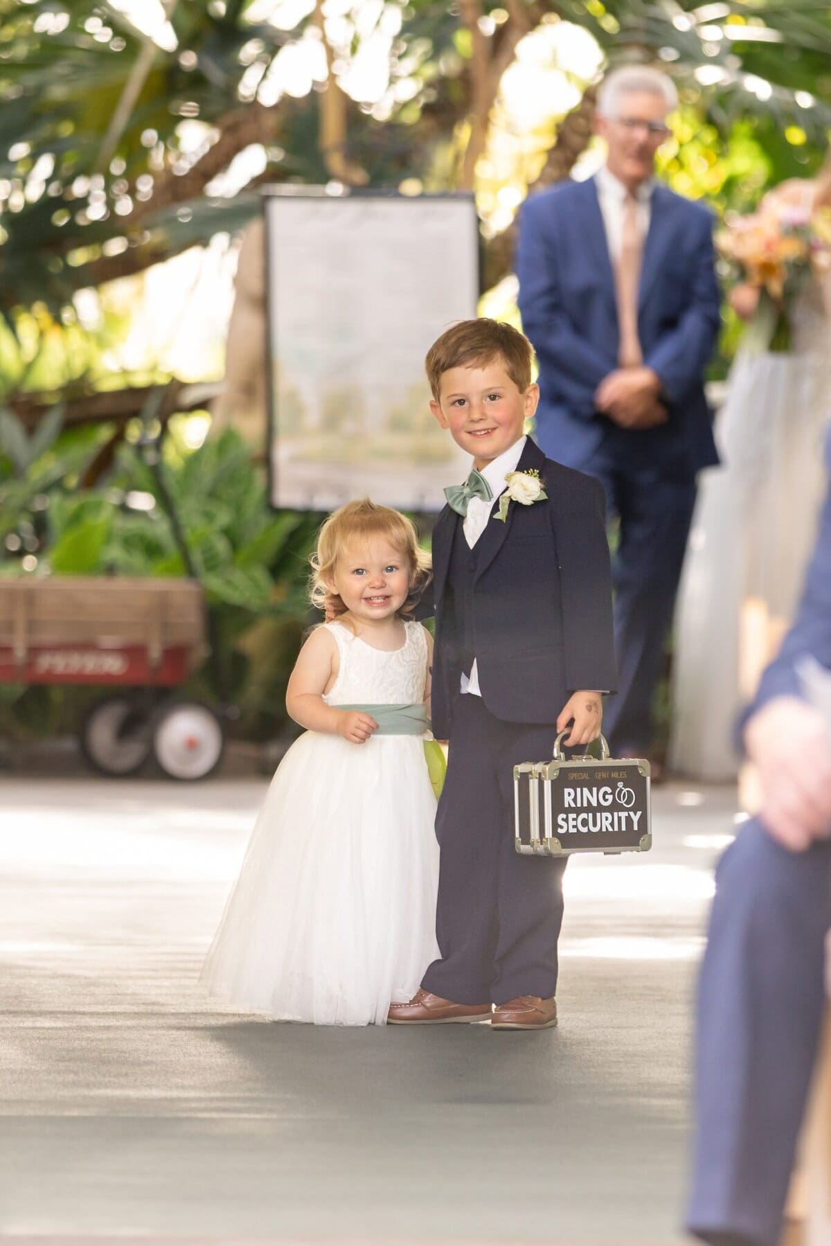 Stunning Wedding at the Roger Williams Botanical Gardens 16 A young boy in a suit holds a "Ring Security" briefcase beside a smiling girl in white at a wedding in the Roger Williams Botanical Gardens.