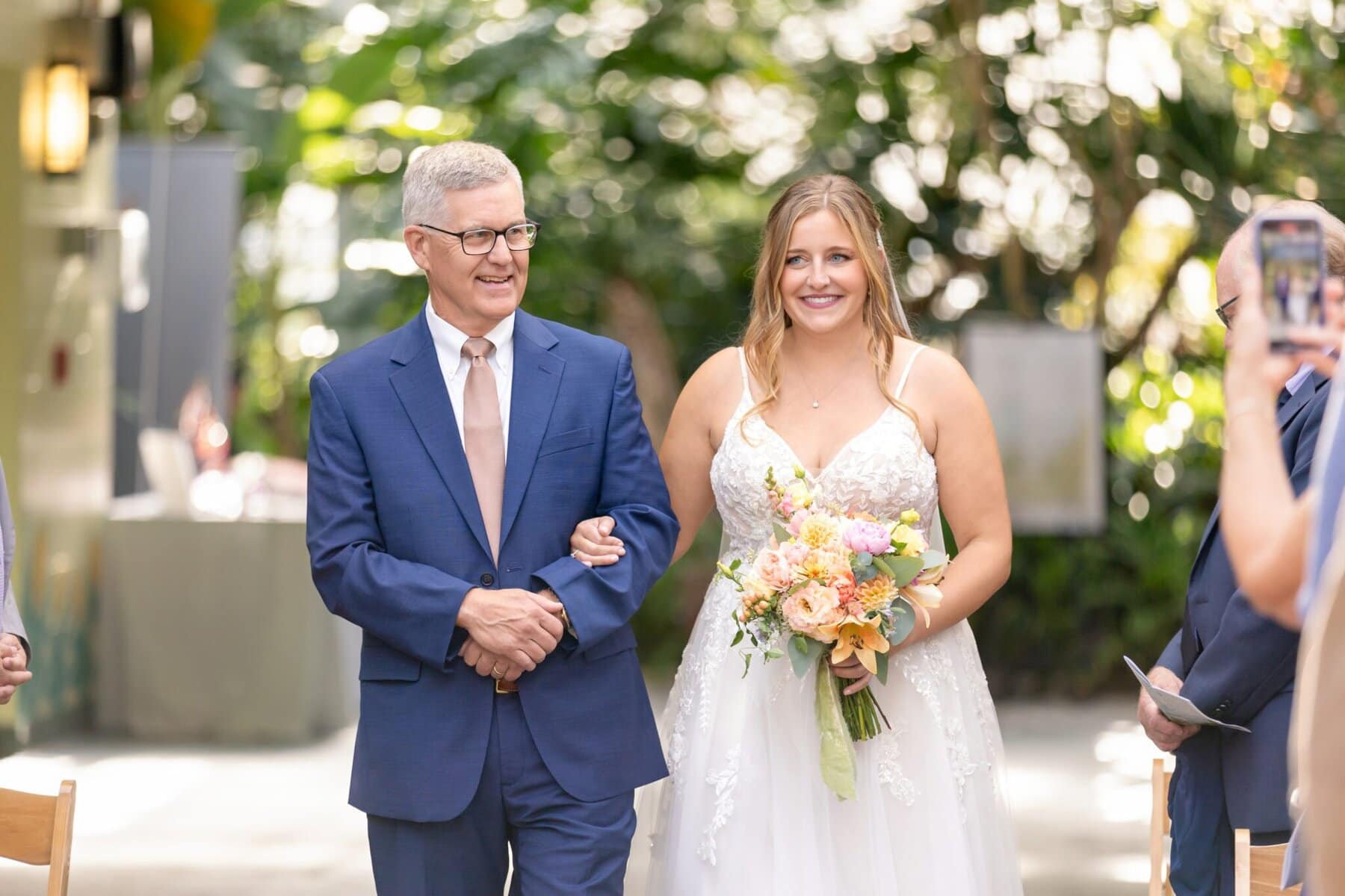 Stunning Wedding at the Roger Williams Botanical Gardens 24 A man in a blue suit walks arm in arm with a bride holding a bouquet at a wedding in Roger Williams Botanical Gardens.