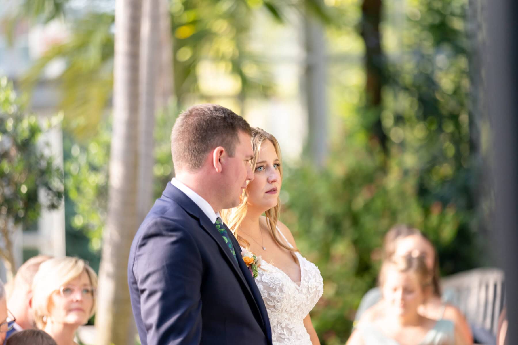 Stunning Wedding at the Roger Williams Botanical Gardens 33 A bride and groom stand together outdoors at Roger Williams Botanical Gardens during their wedding, with guests seated nearby.