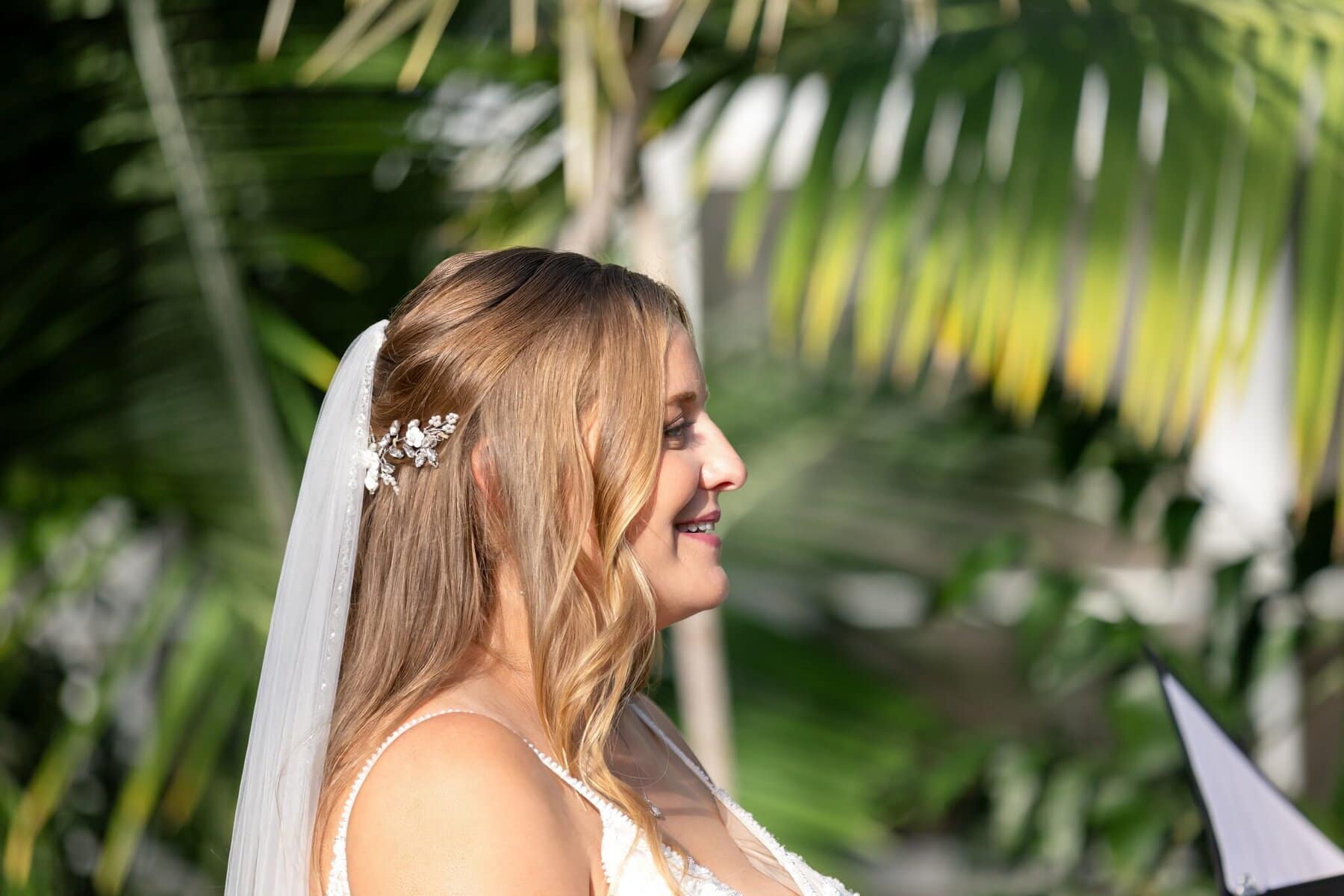 Stunning Wedding at the Roger Williams Botanical Gardens 29 A bride with long blonde hair and a veil stands outdoors, smiling, framed by palm leaves at Roger Williams Botanical Gardens.