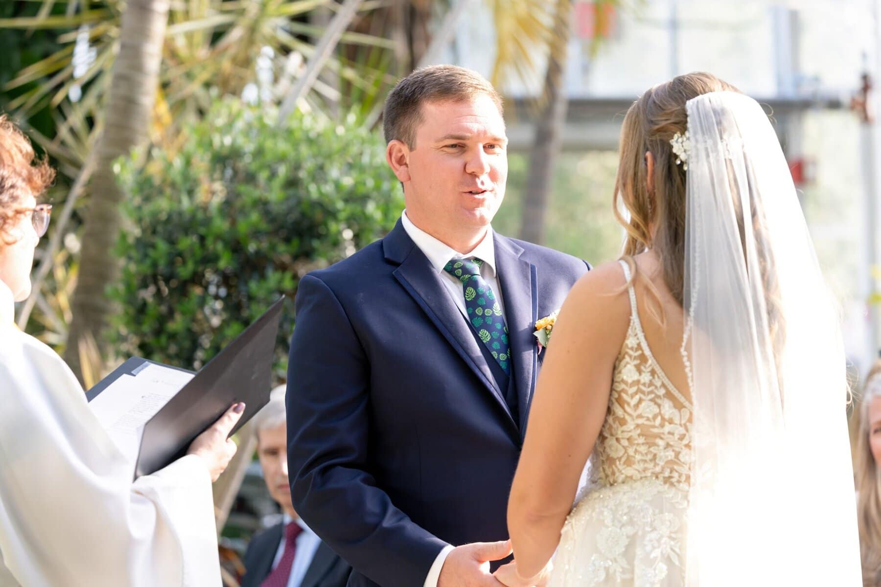 Stunning Wedding at the Roger Williams Botanical Gardens 28 A groom and bride stand facing each other during a wedding at Roger Williams Botanical Gardens, with an officiant holding papers nearby.