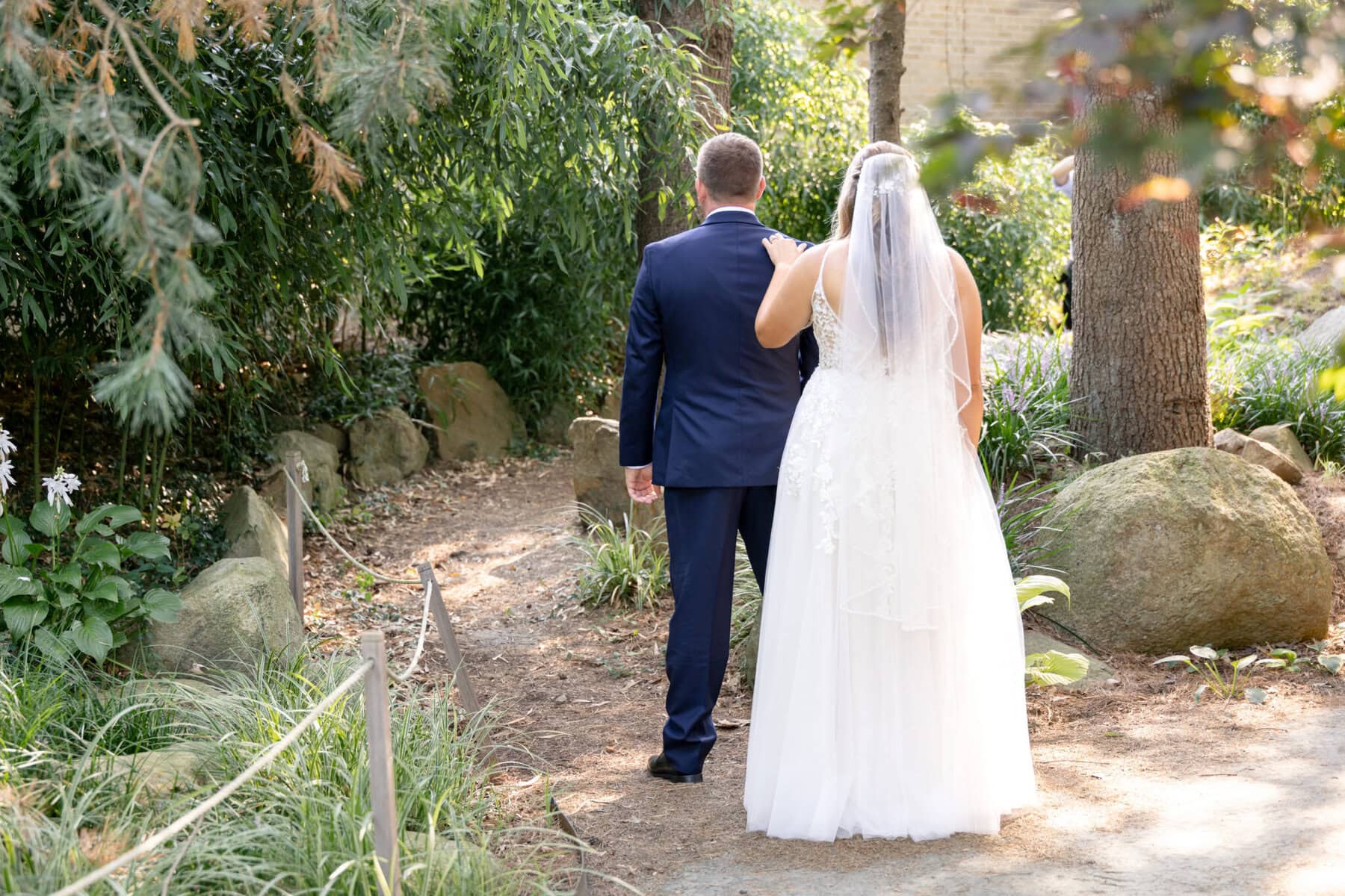 Stunning Wedding at the Roger Williams Botanical Gardens 7 A bride and groom stand side by side on a garden path at Roger Williams Botanical Gardens, surrounded by greenery and rocks.