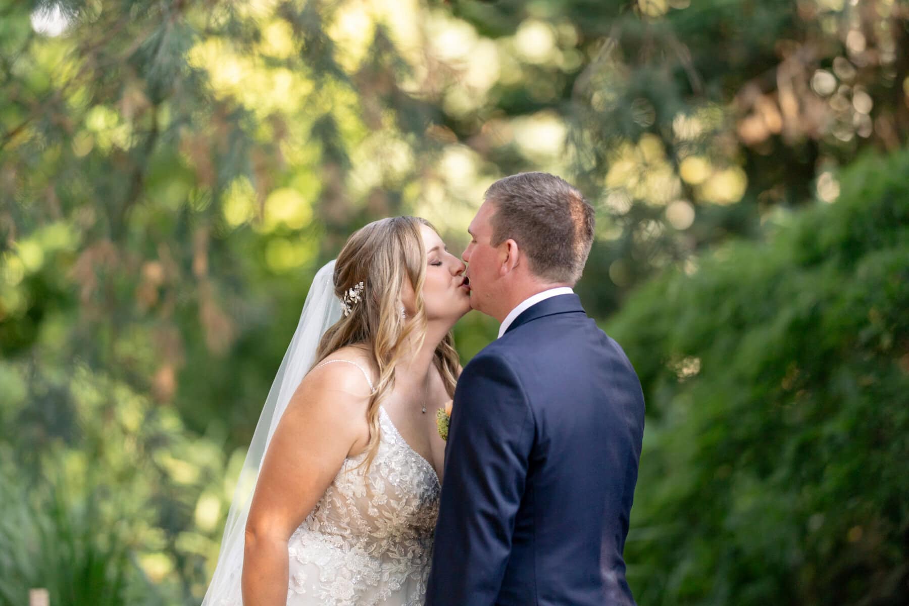Stunning Wedding at the Roger Williams Botanical Gardens 10 A bride and groom share a kiss outdoors at Roger Williams Botanical Gardens, surrounded by lush foliage and soft sunlight.