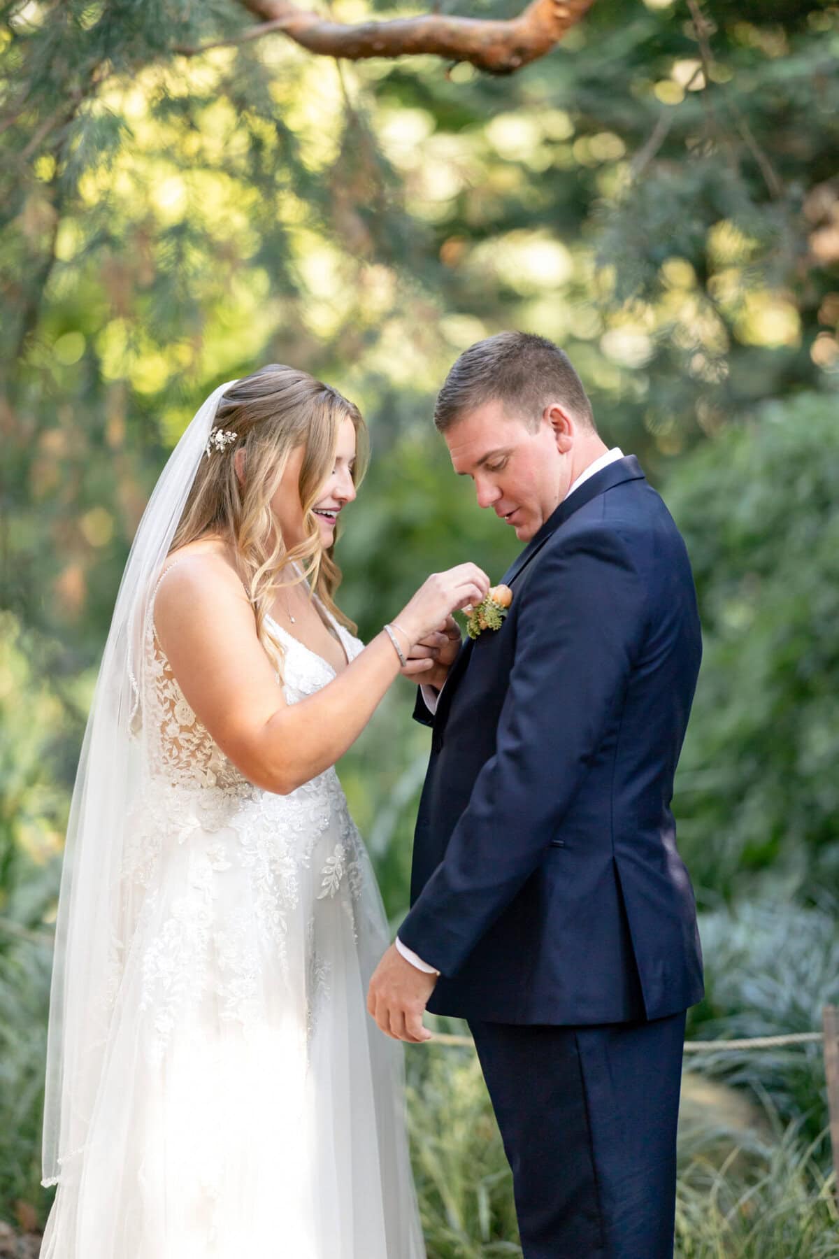 Stunning Wedding at the Roger Williams Botanical Gardens 11 A bride in a white dress adjusts the groom’s boutonniere outdoors during their wedding at Roger Williams Botanical Gardens.