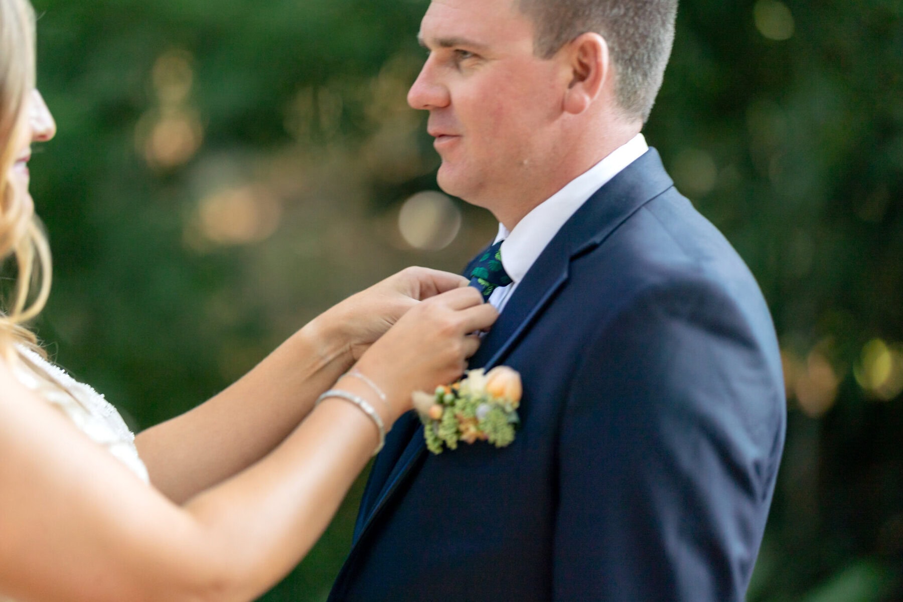 Stunning Wedding at the Roger Williams Botanical Gardens 13 A person in a suit stands as another adjusts their tie, with greenery and blurred trees of Roger Williams Botanical Gardens in the background.