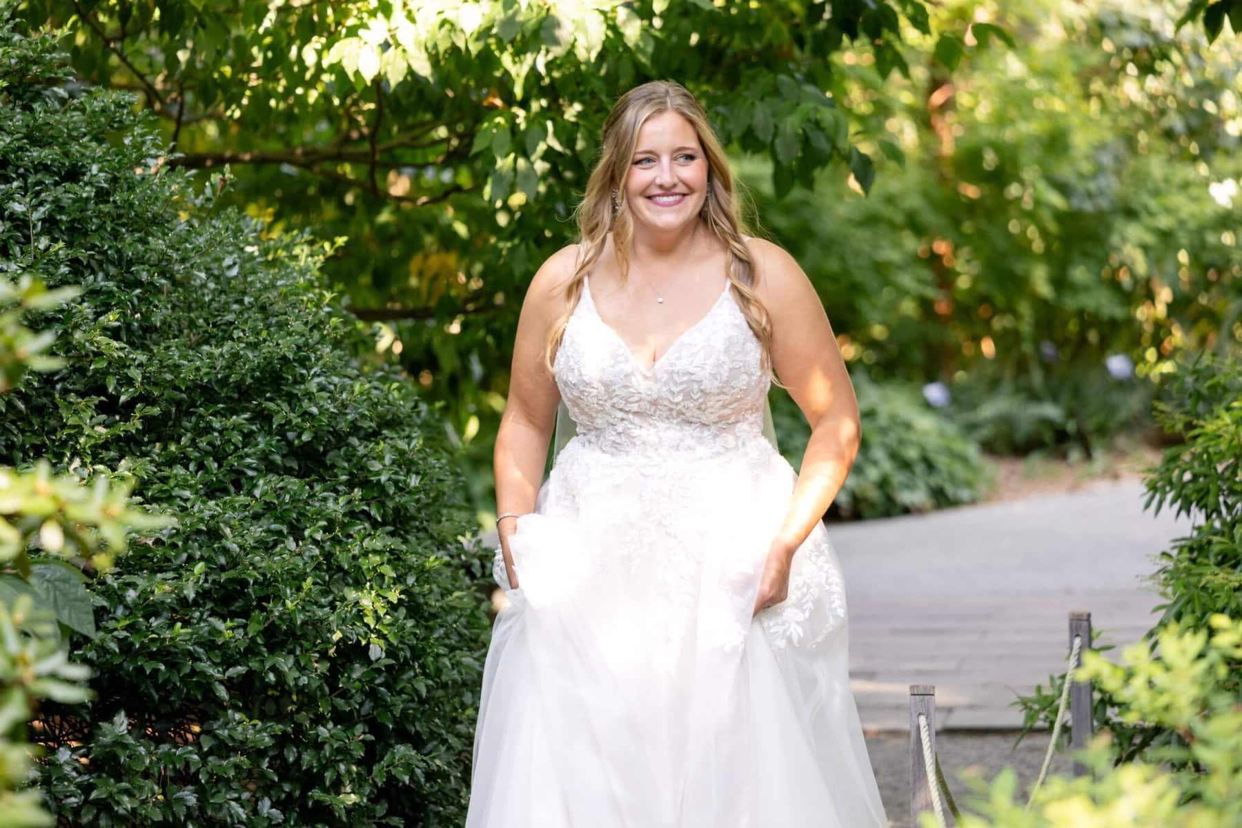 Stunning Wedding at the Roger Williams Botanical Gardens 4 A woman in a white wedding dress stands outdoors on a sunny day at the Roger Williams Botanical Gardens, surrounded by lush greenery.