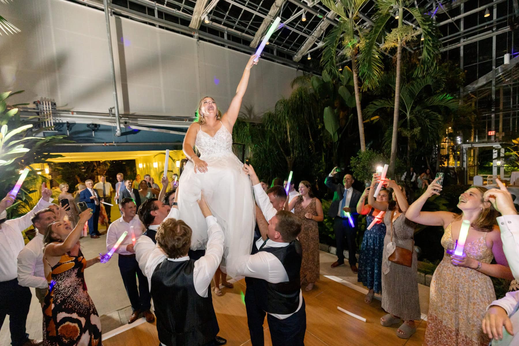 Stunning Wedding at the Roger Williams Botanical Gardens 91 At a wedding in Roger Williams Botanical Gardens, a bride is lifted on a chair as guests surround her with glowing light sticks.