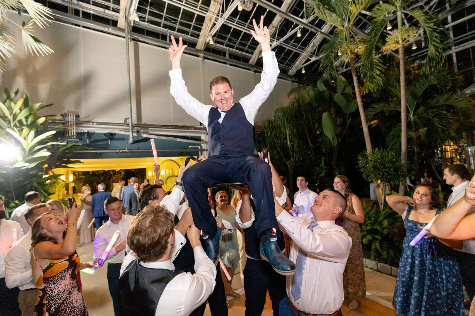 Stunning Wedding at the Roger Williams Botanical Gardens 92 A man in a suit is lifted in the air at a wedding in Roger Williams Botanical Gardens, smiling and raising both hands in victory.