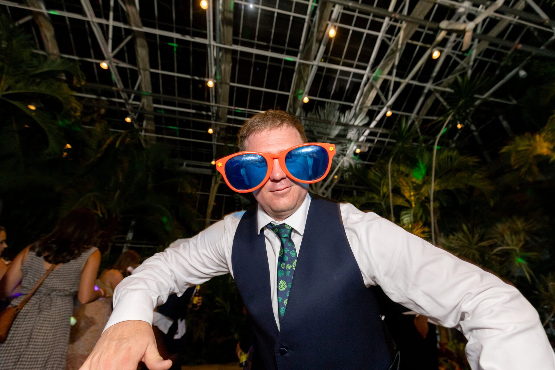 Stunning Wedding at the Roger Williams Botanical Gardens 93 A man in a suit and tie sports oversized orange sunglasses at a wedding inside Roger Williams Botanical Gardens.