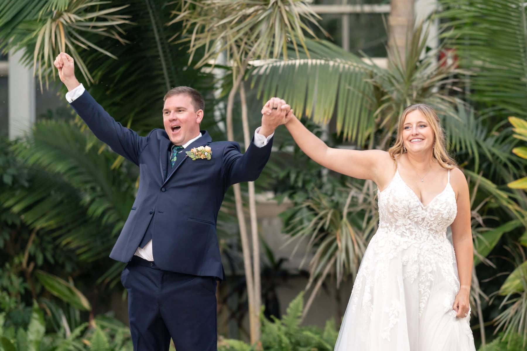 Stunning Wedding at the Roger Williams Botanical Gardens 64 A bride and groom stand outside holding hands, smiling; the groom raises his arm in celebration at Roger Williams Botanical Gardens.