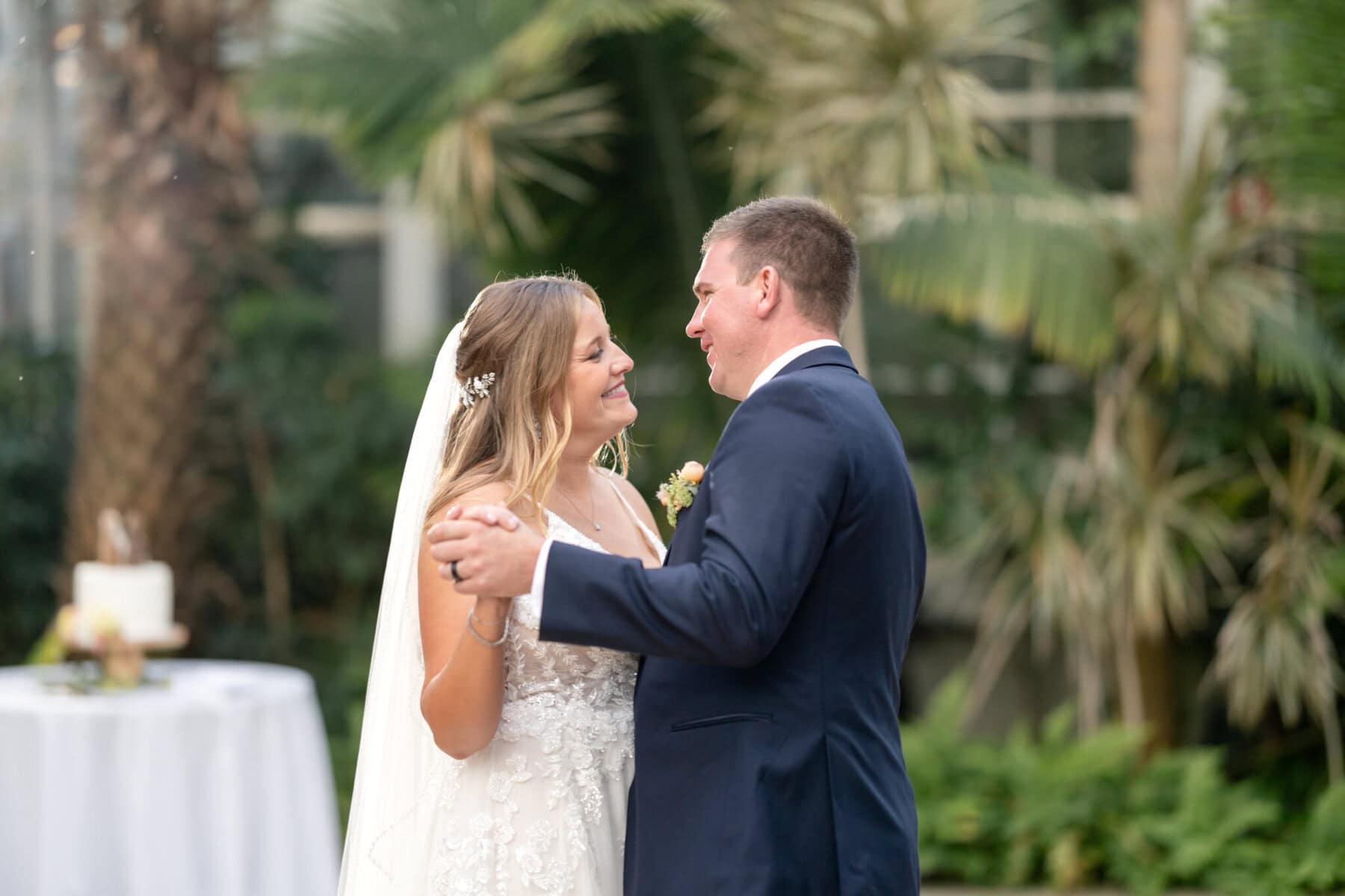 Stunning Wedding at the Roger Williams Botanical Gardens 65 A bride and groom smile at each other while dancing outdoors during their Roger Williams Botanical Gardens wedding, with cake and palms behind them.