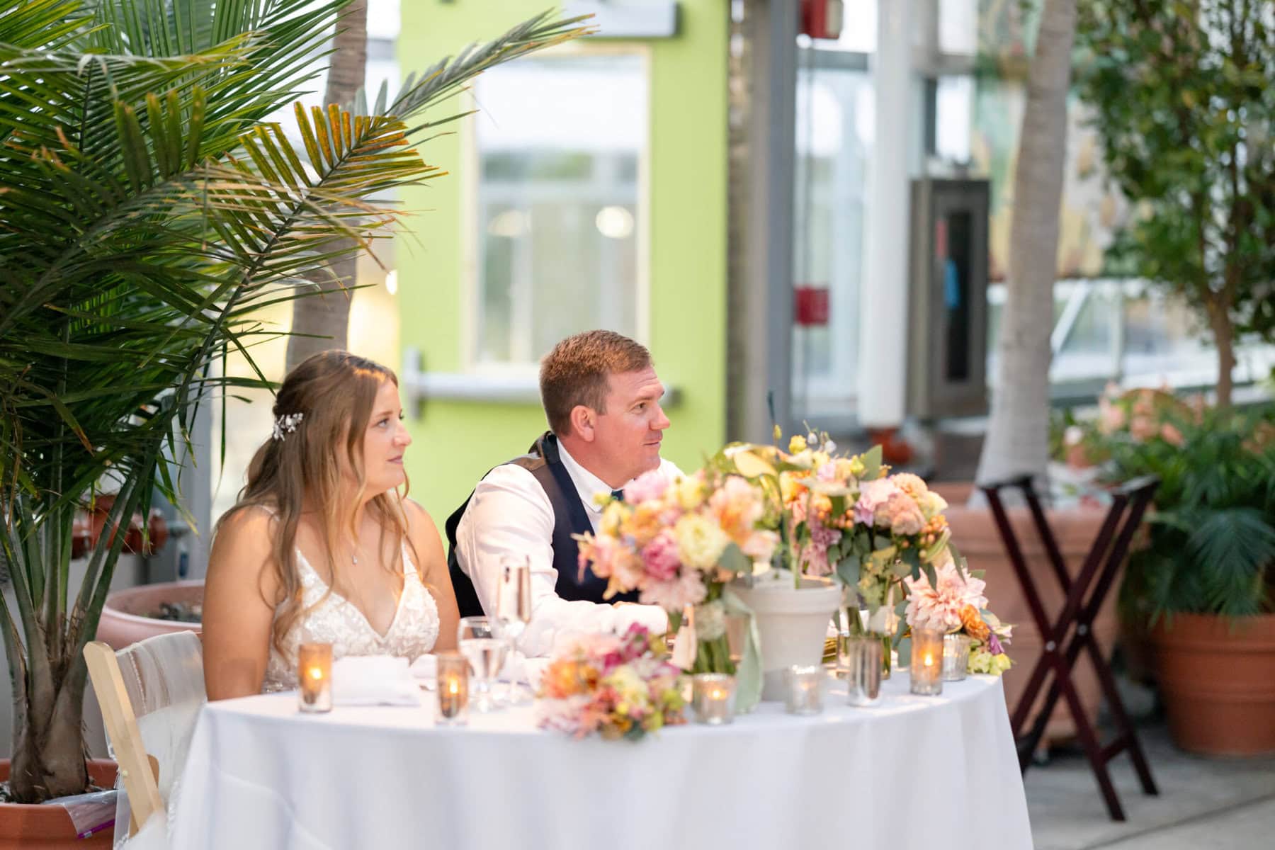 Stunning Wedding at the Roger Williams Botanical Gardens 71 A bride and groom sit at a round table adorned with flowers and candles in the bright Roger Williams Botanical Gardens wedding venue.