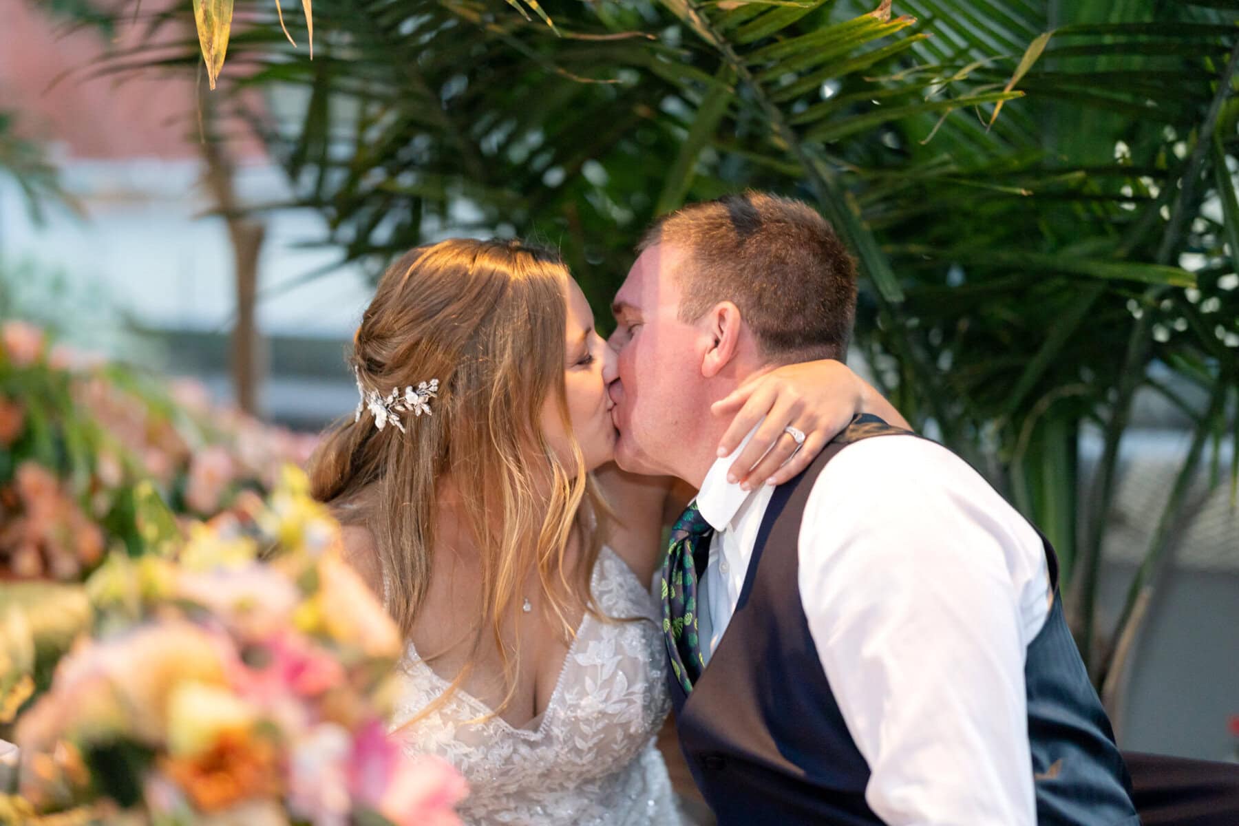 Stunning Wedding at the Roger Williams Botanical Gardens 73 A bride and groom kiss while seated at their wedding, surrounded by greenery and colorful flowers in Roger Williams Botanical Gardens.